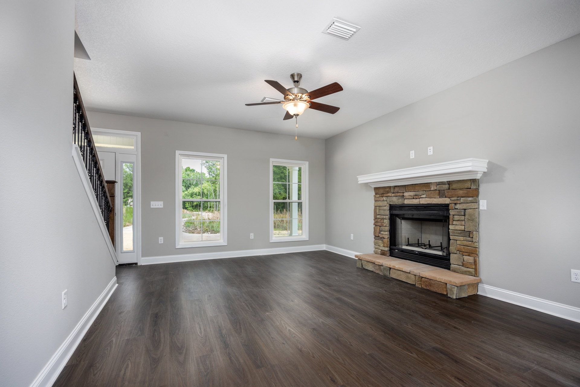Living room with hardwood flooring, white-framed window showing trees outside, glass-door fireplace set in a neutral wall, and ceiling fan with light fixture.