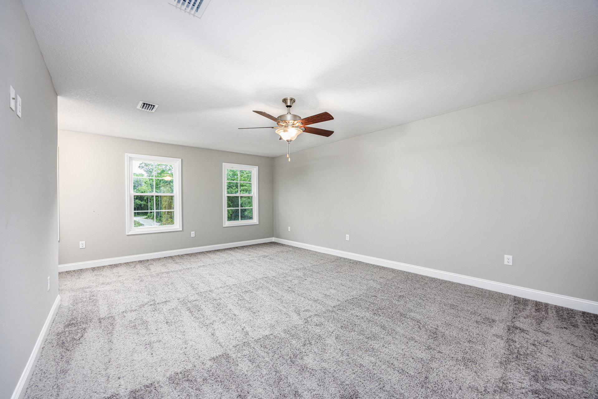 Bedroom with grey carpet, white walls, ceiling fan with light fixture, large windows showing green trees outside, simple crown molding