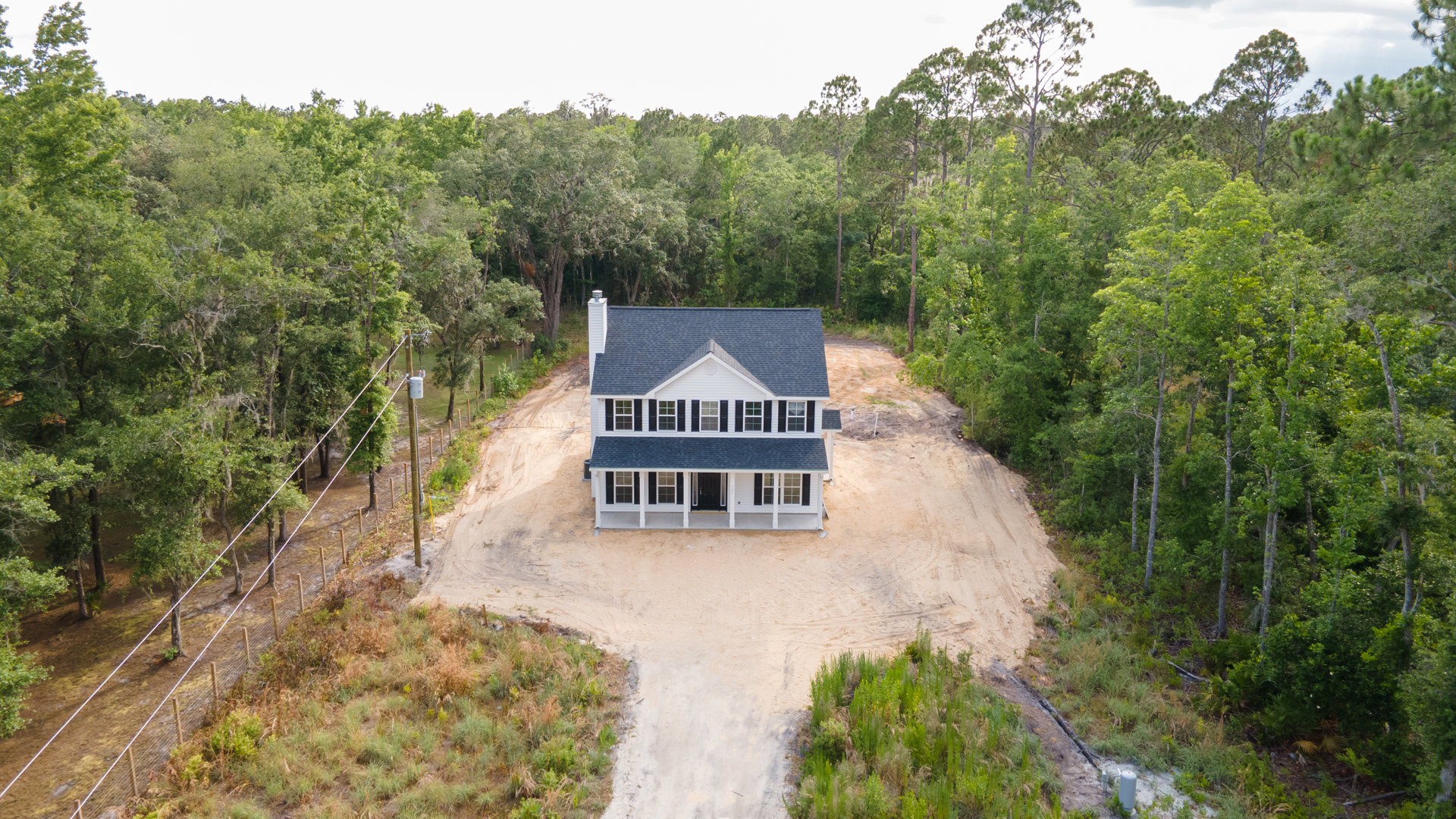 Modern house with black roof and large windows, situated on a dirt road and surrounded by dense trees and greenery, power lines visible along the road