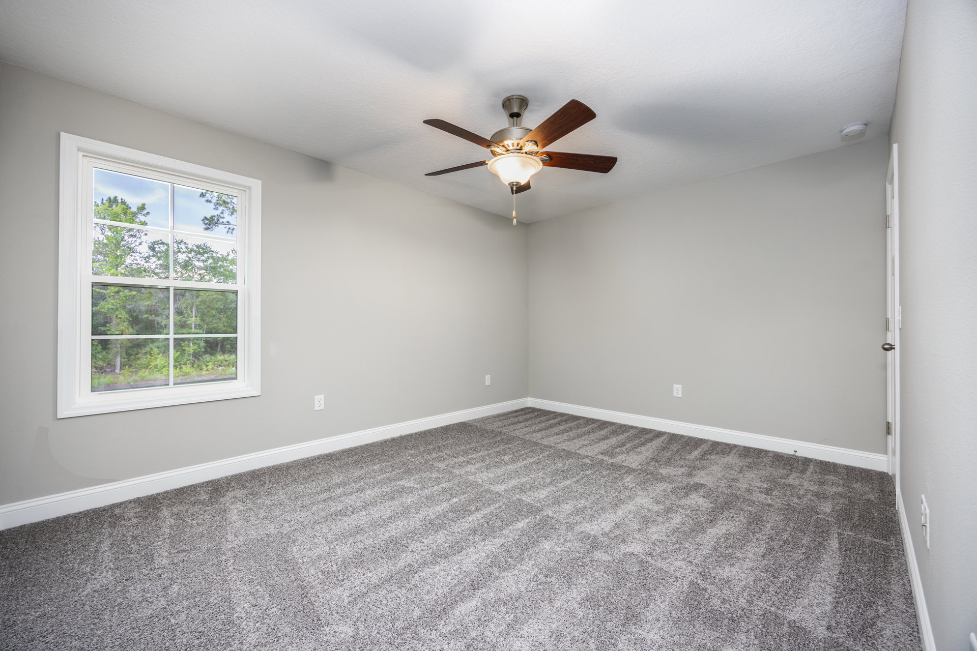 Ceiling fan with light fixture above grey carpeted floor, white walls, large window showing trees outside