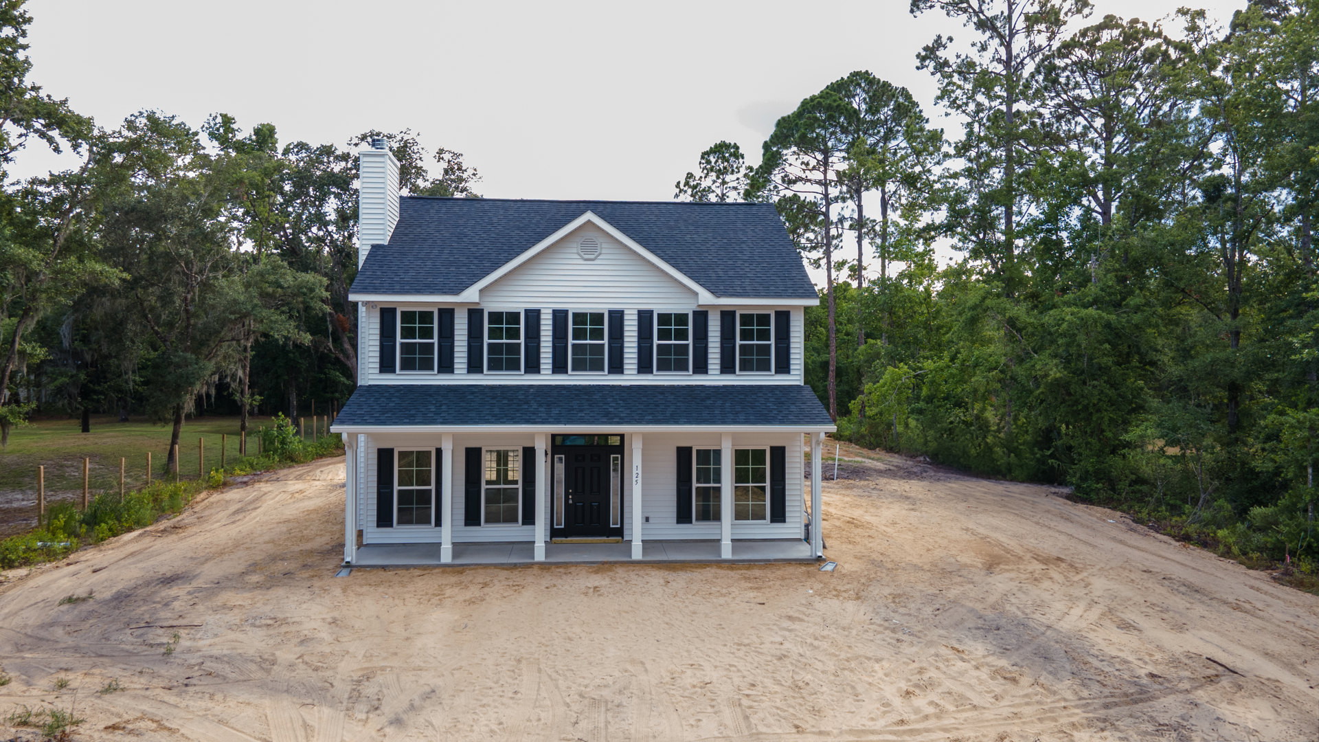 White house with black front door, glass panel, and white pillars, surrounded by trees, dirt road in foreground, large windows visible on exterior