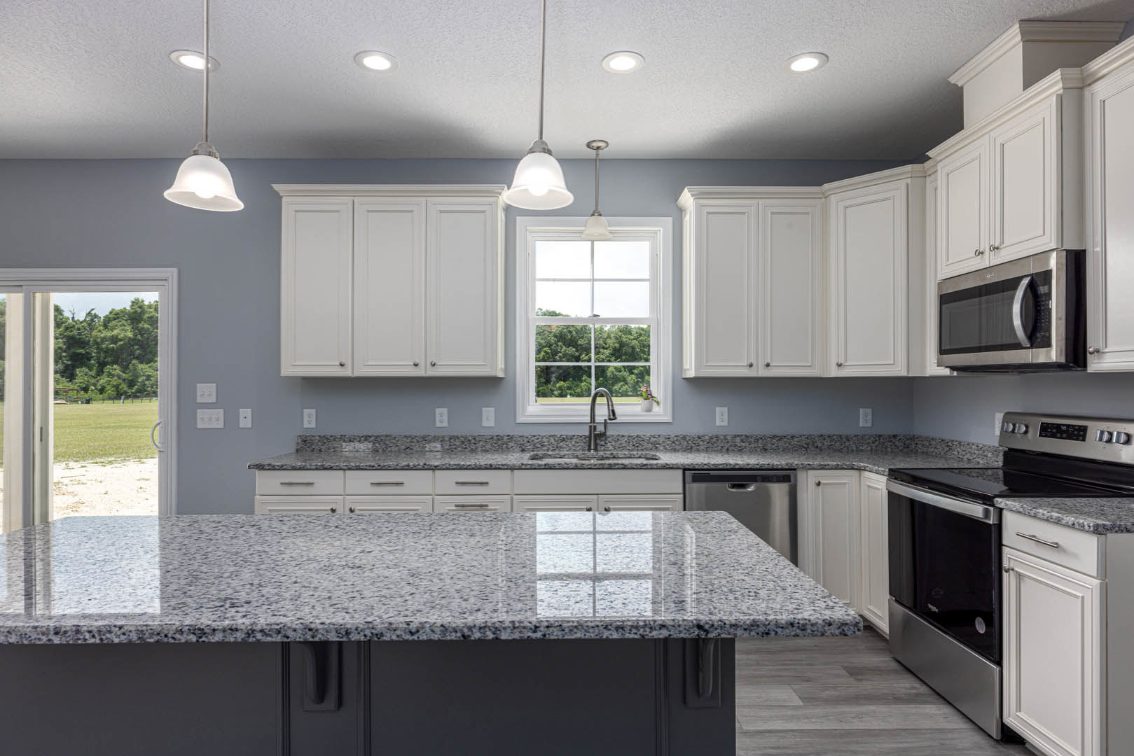 Granite countertops, white shaker cabinets, stainless steel microwave and oven, undermount sink beneath window with view of trees, chrome faucet, neutral tile backsplash