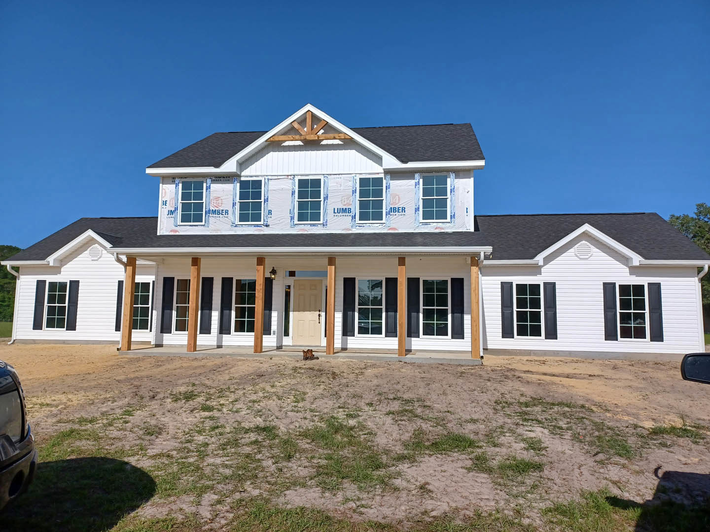 Modern home under construction with exposed framing, unfinished exterior walls, and a dirt yard featuring scattered construction materials; black window shutters and roof visible