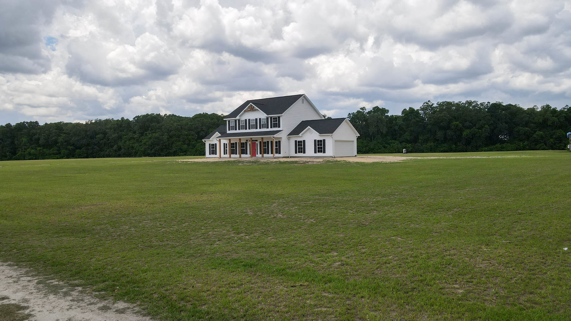 White farmhouse with black shutters and red door set behind expansive green field, surrounded by trees under a cloudy sky