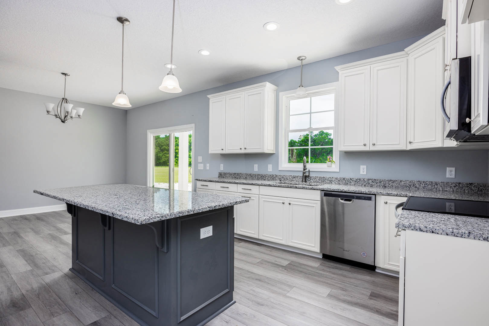 Granite countertops and white cabinets in a kitchen with a marble-topped island, stainless steel dishwasher, white electrical outlet, and a window overlooking trees