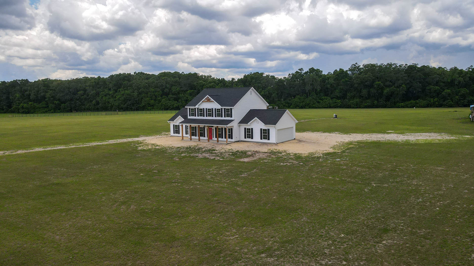 White house with black roof, red door, and black shutters set in a grassy field with trees and cloudy sky in the background