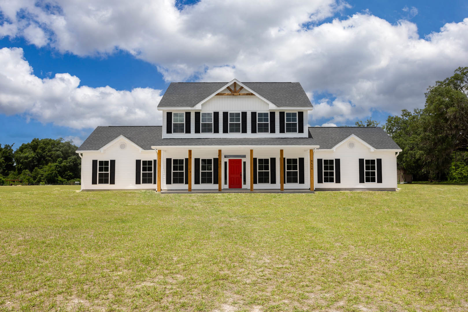 White two-story house with black-framed windows, red front door with black handles, covered porch, manicured lawn, and mature trees under a partly cloudy sky