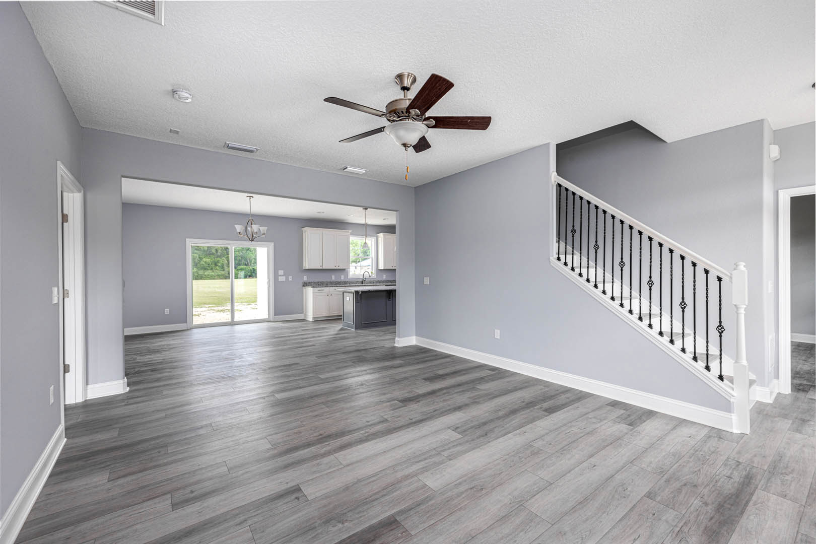 Open living area with wood flooring, white cabinets along one wall, ceiling fan with light fixture, staircase featuring wooden steps and white railing, large window overlooking