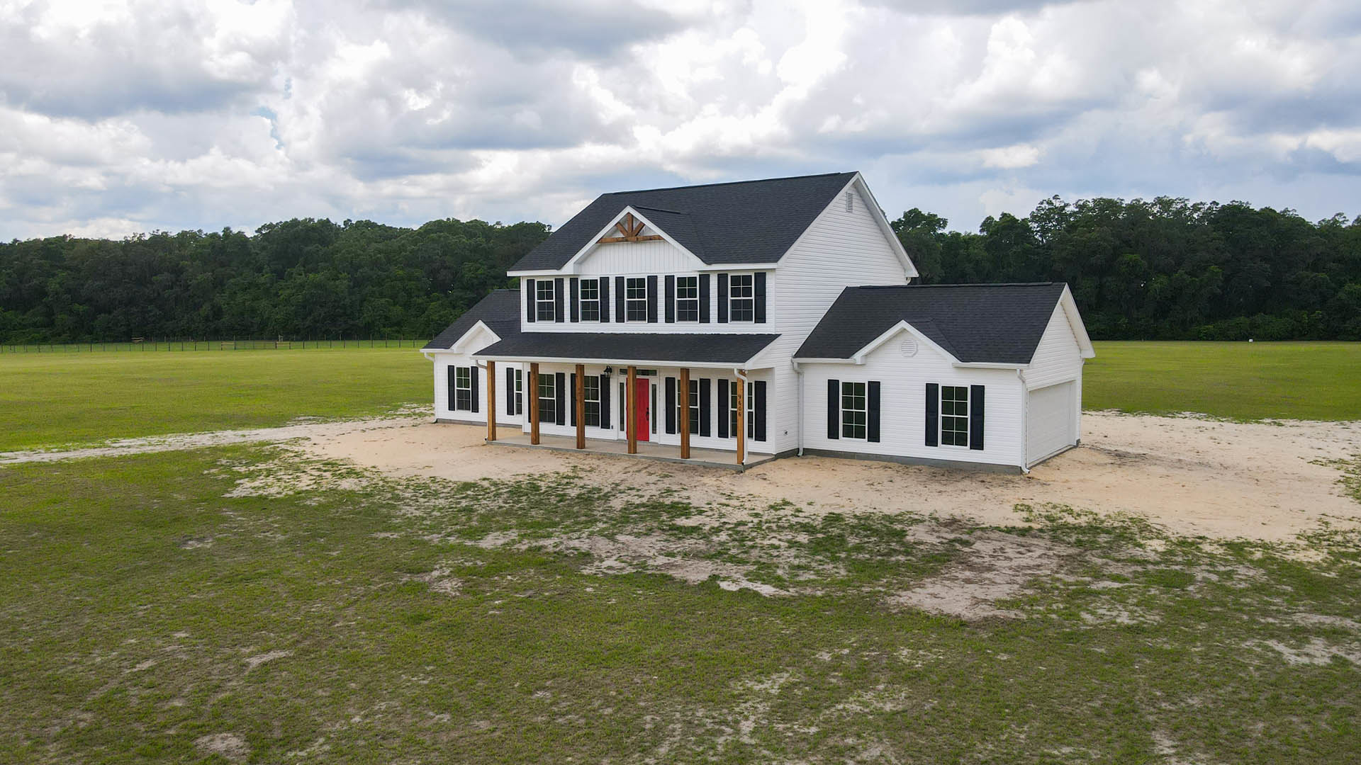 White two-story house with black shutters, expansive covered front porch, manicured lawn, dirt landscaping, and cloudy sky overhead