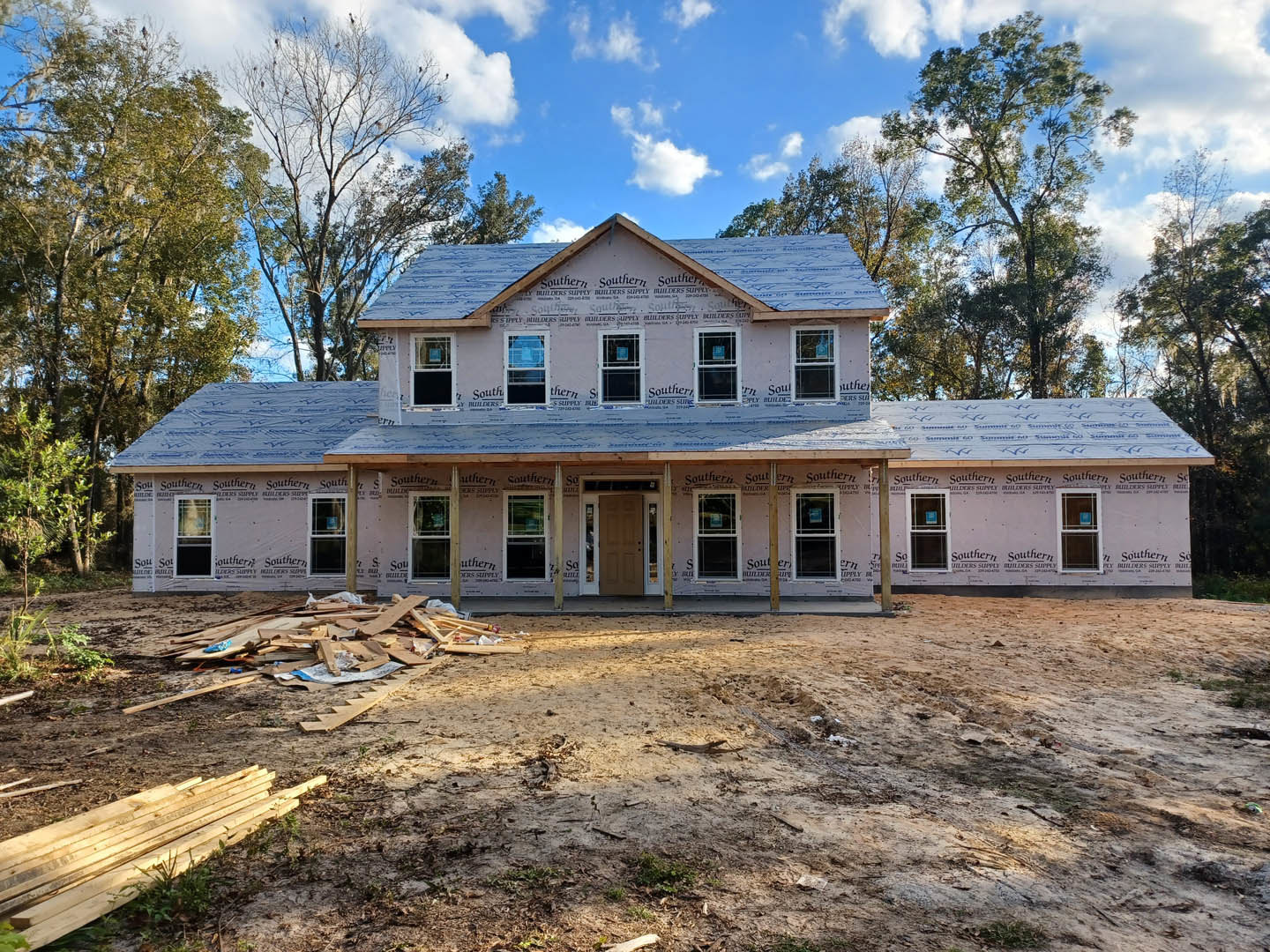 Partially built farmhouse with exposed wooden framing, covered porch, blue glass window, dirt yard, and surrounding trees