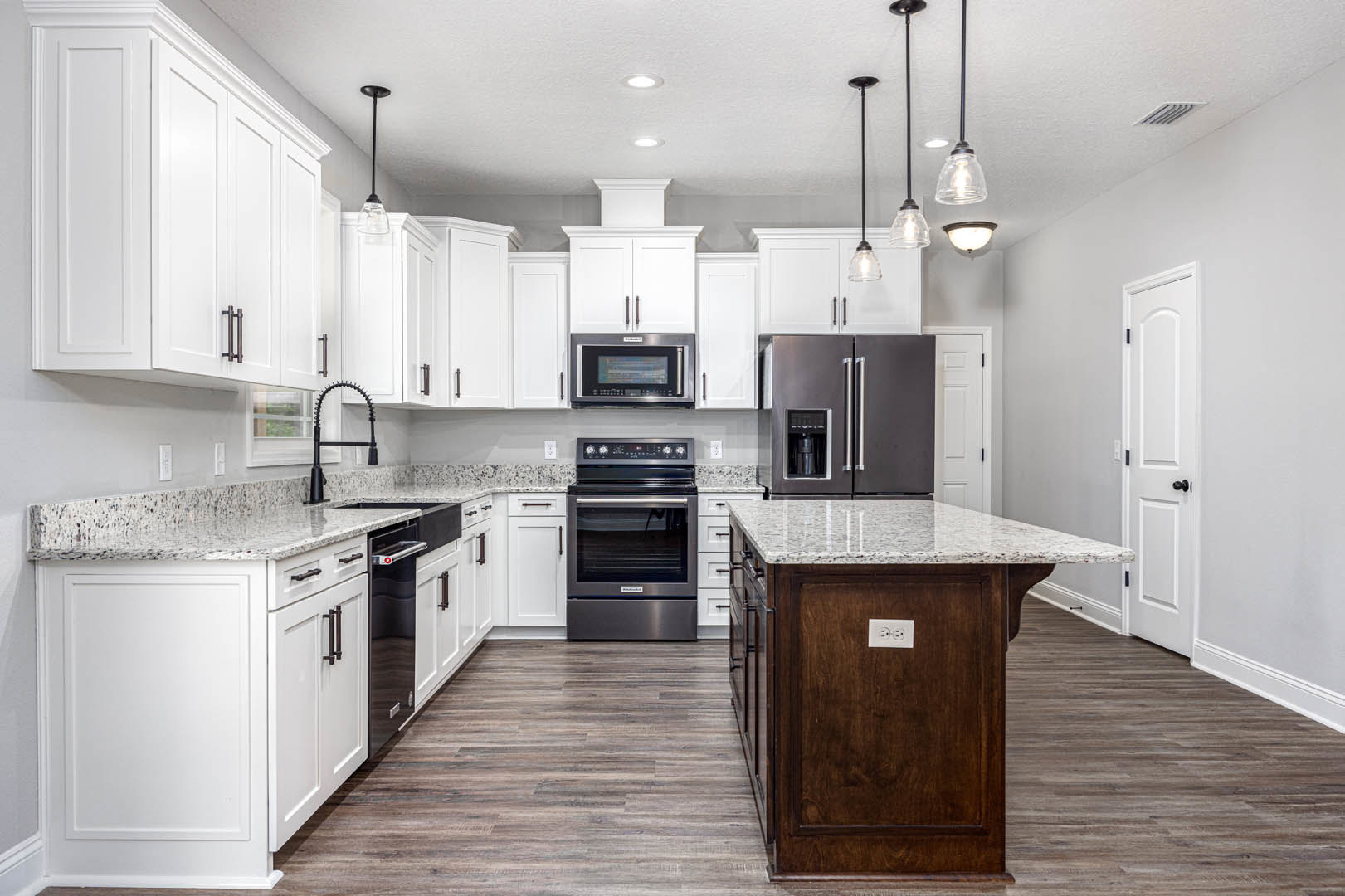 White kitchen with granite countertops, stainless steel oven, microwave, and refrigerator, marble-topped island, pendant light, and wood flooring.