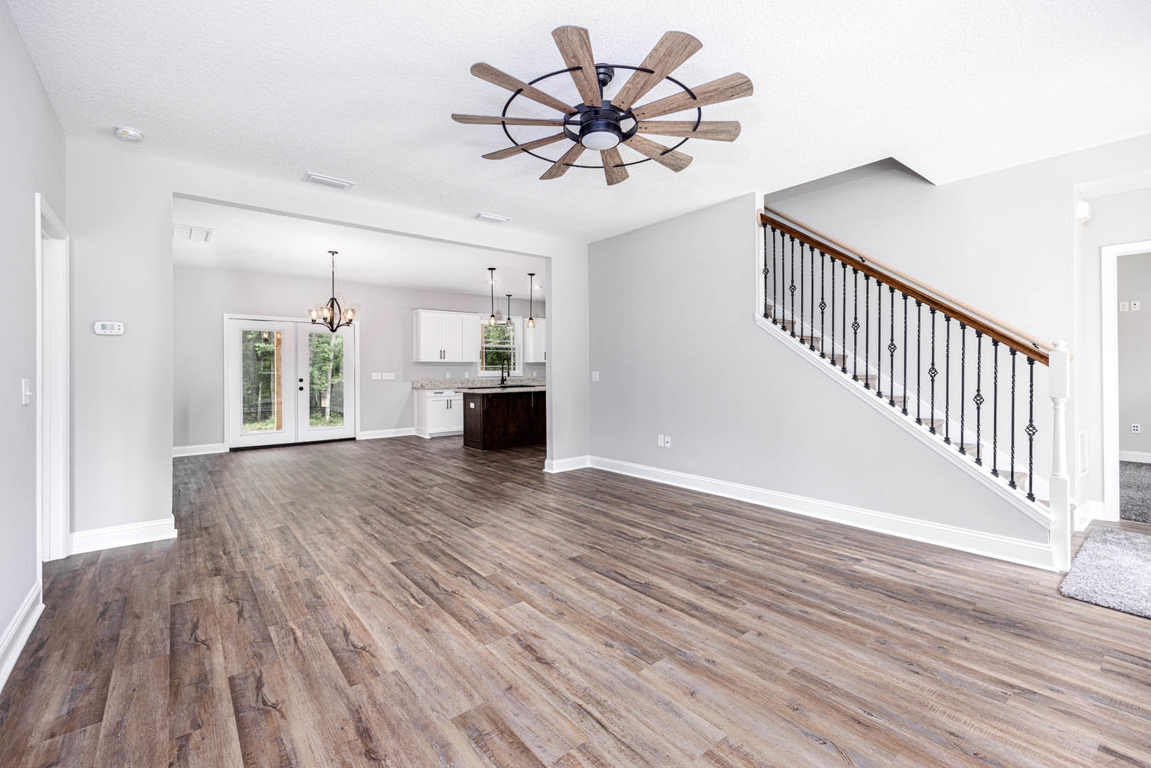 Open living area with wood flooring, ceiling fan with light, staircase featuring metal railing, white double doors with glass panes, and brown console table topped with marble