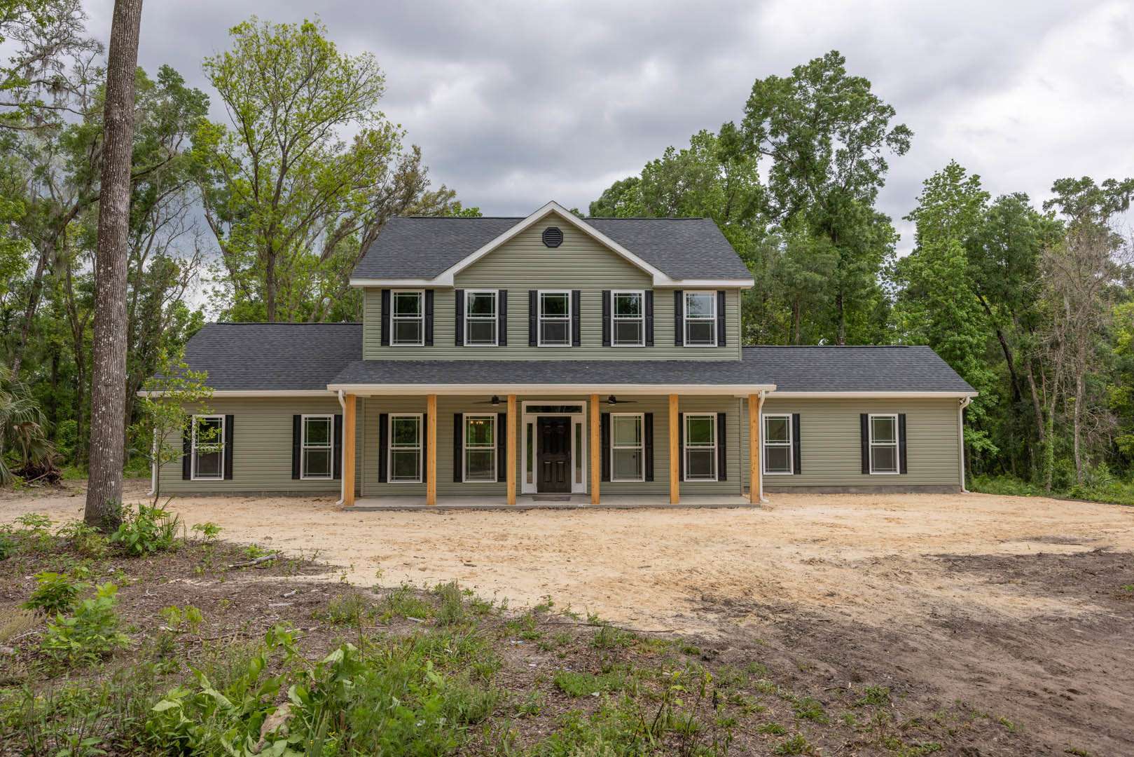 Two-story home with white siding, black front door, large windows, spacious concrete driveway, manicured lawn, and mature trees under a partly cloudy sky