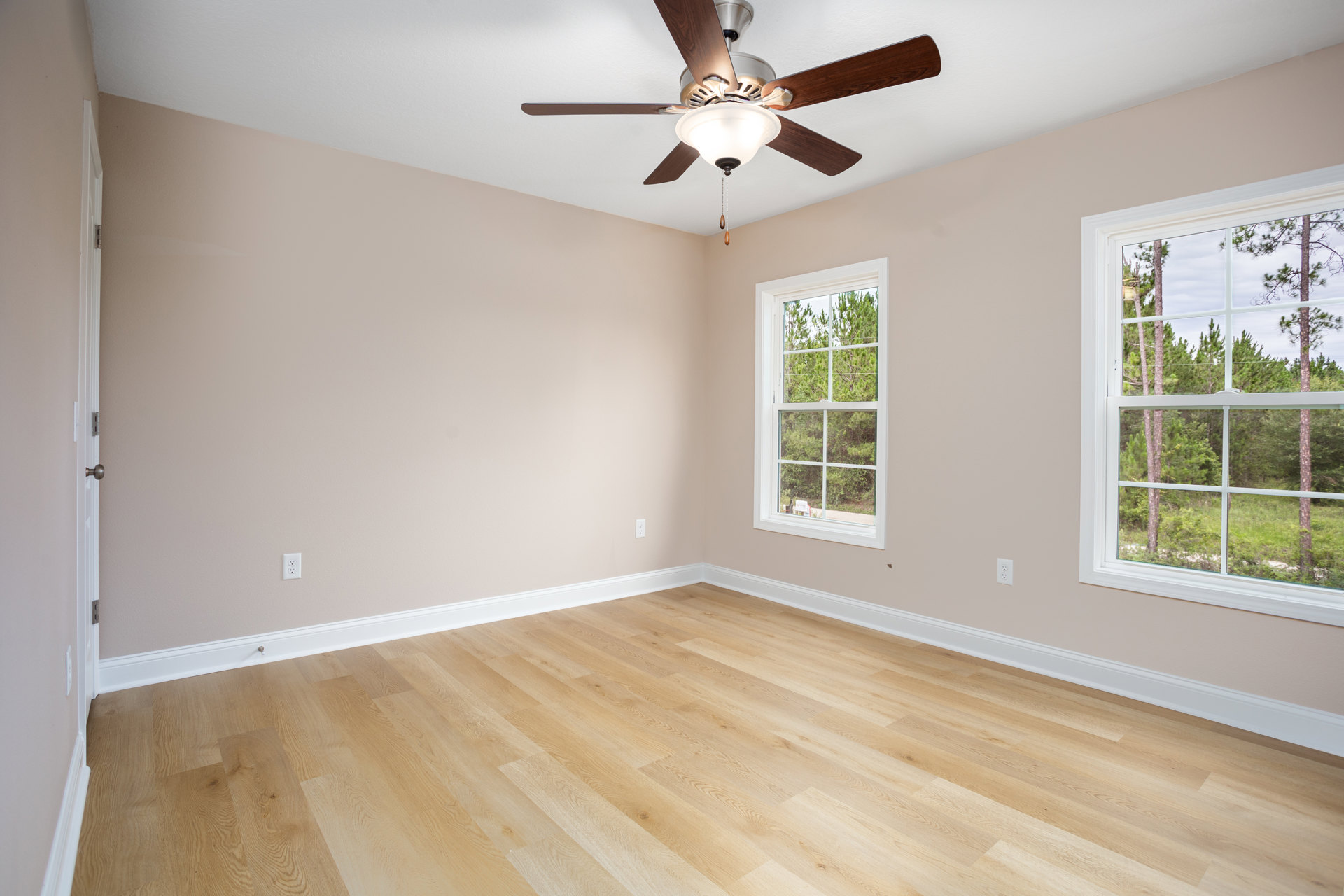 Ceiling fan with light fixture, large window overlooking trees, wood flooring with white baseboard trim, neutral walls and ceiling