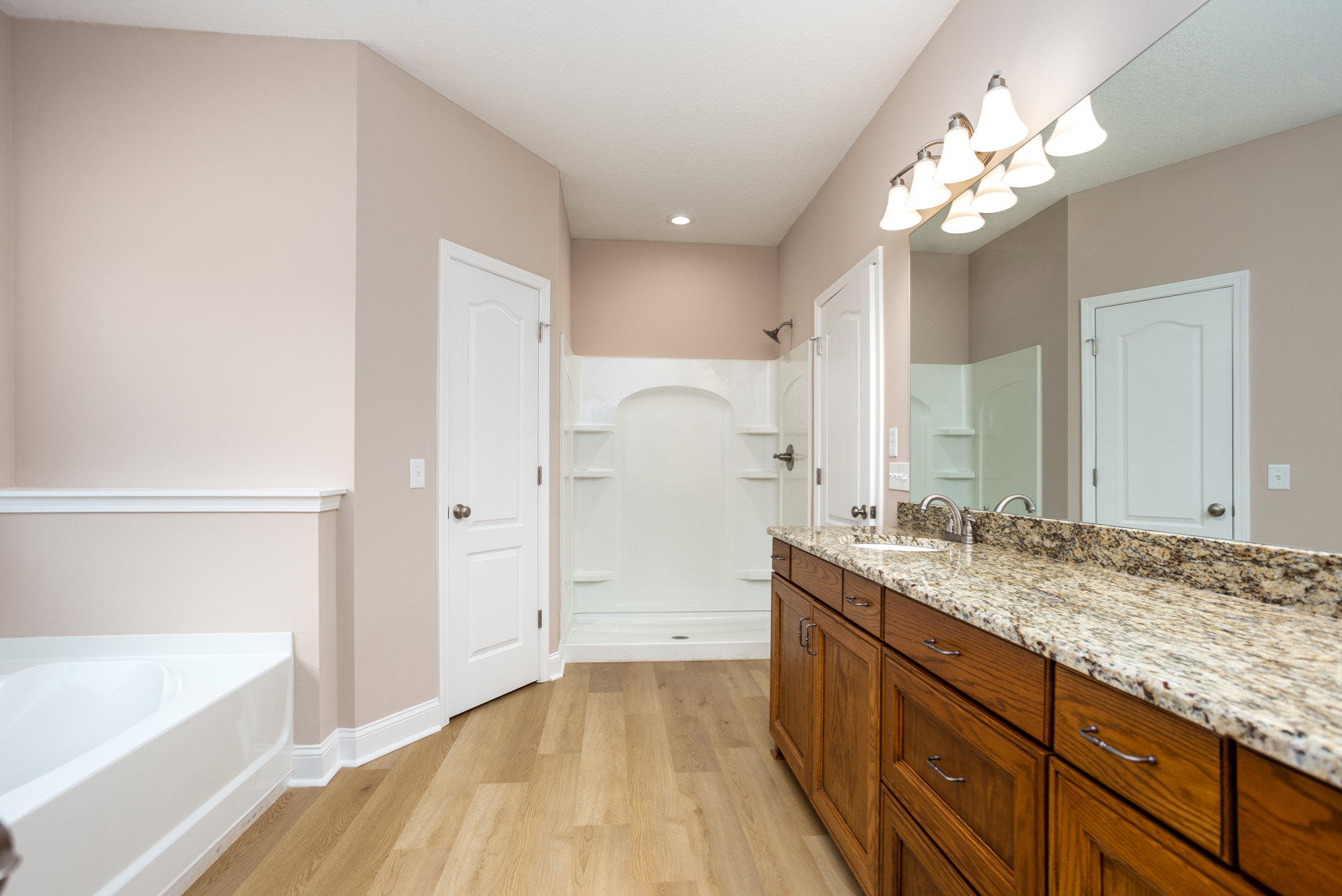 Bathroom with marble countertop, undermount sink, large mirror framed by wall lights, freestanding white bathtub, white paneled door with chrome doorknob, light tile flooring, and