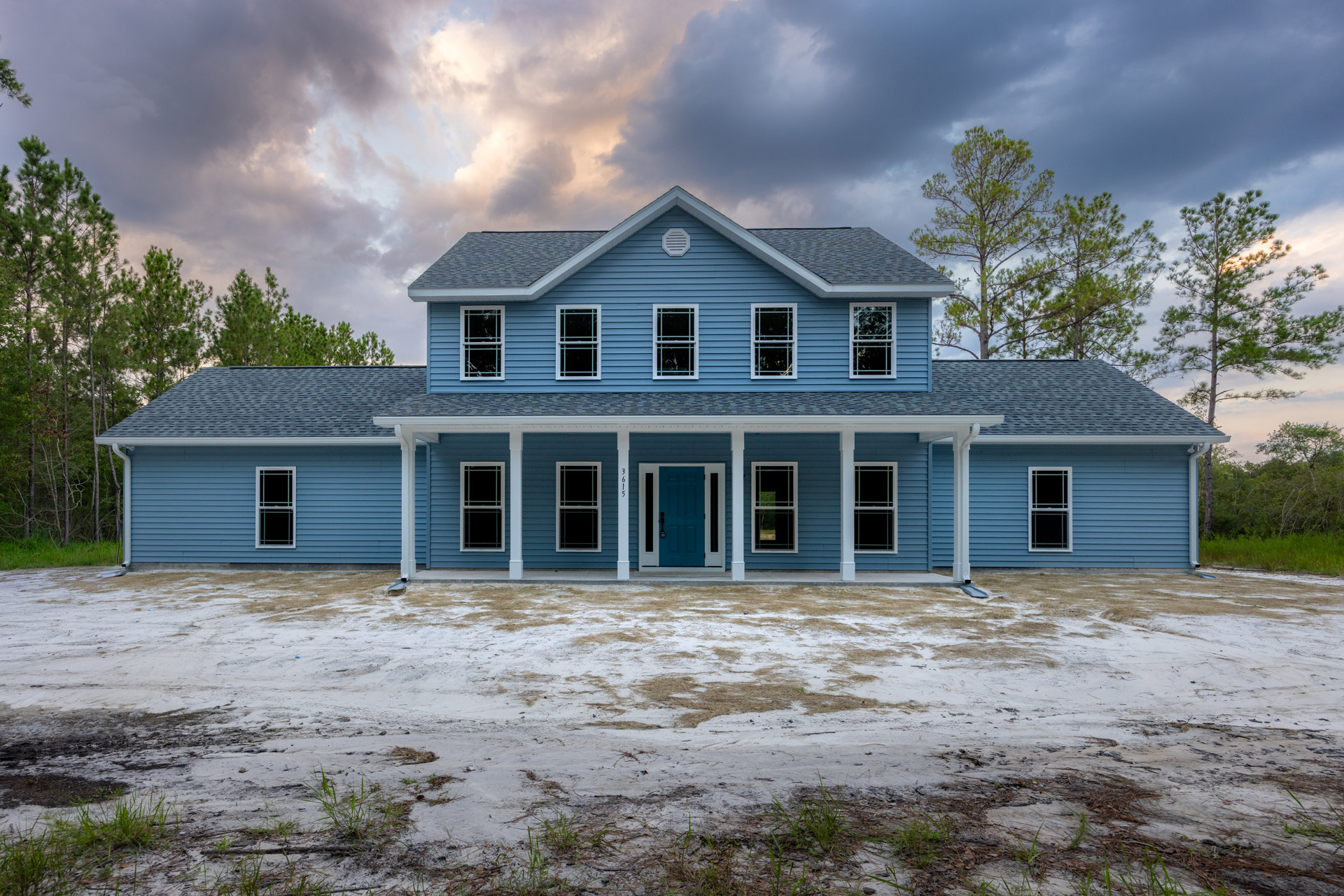 Blue two-story house with white pillars, white trim windows, dirt yard in front, broken glass in one window, cloudy sky overhead, trees in background.