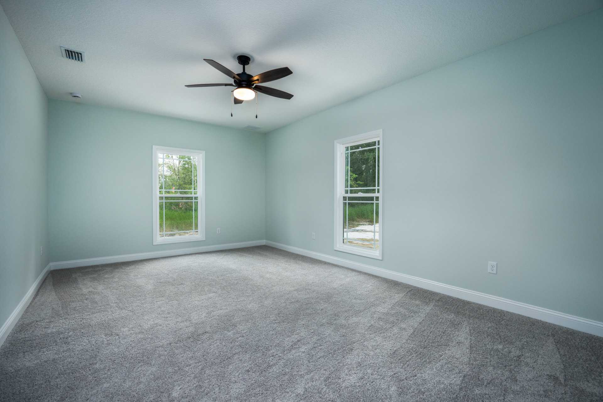 Carpeted bedroom with white walls, ceiling fan with light, large windows showing forest view and American flag outside