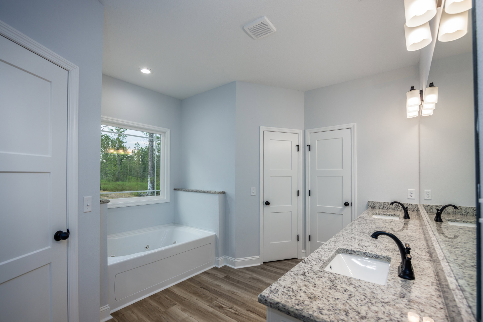 Bathroom featuring a marble countertop with integrated sink, freestanding bathtub, white cabinetry, black door hardware, window overlooking trees, and modern light fixture.