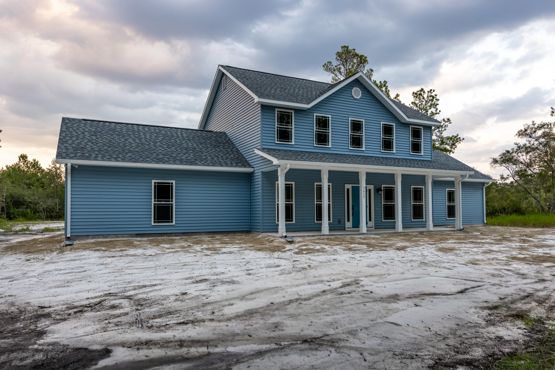 Blue siding house with expansive white porch, white door, multiple windows including one with broken glass and another with black screen, white window frames, dirt ground in front