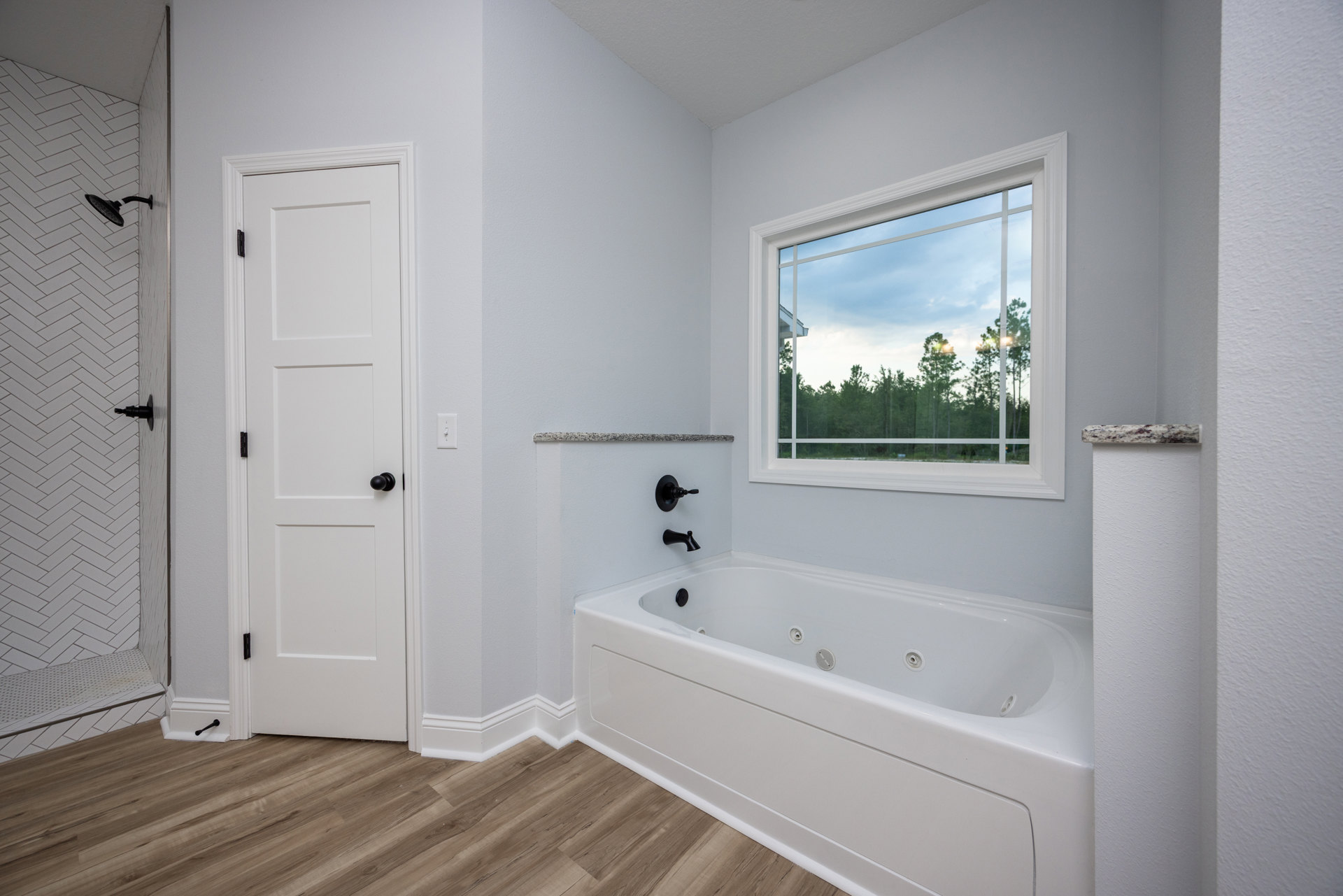 White bathtub with black faucet and spout set against a white tile wall, window overlooking trees, white door with black knob, shower head mounted on tiled wall.
