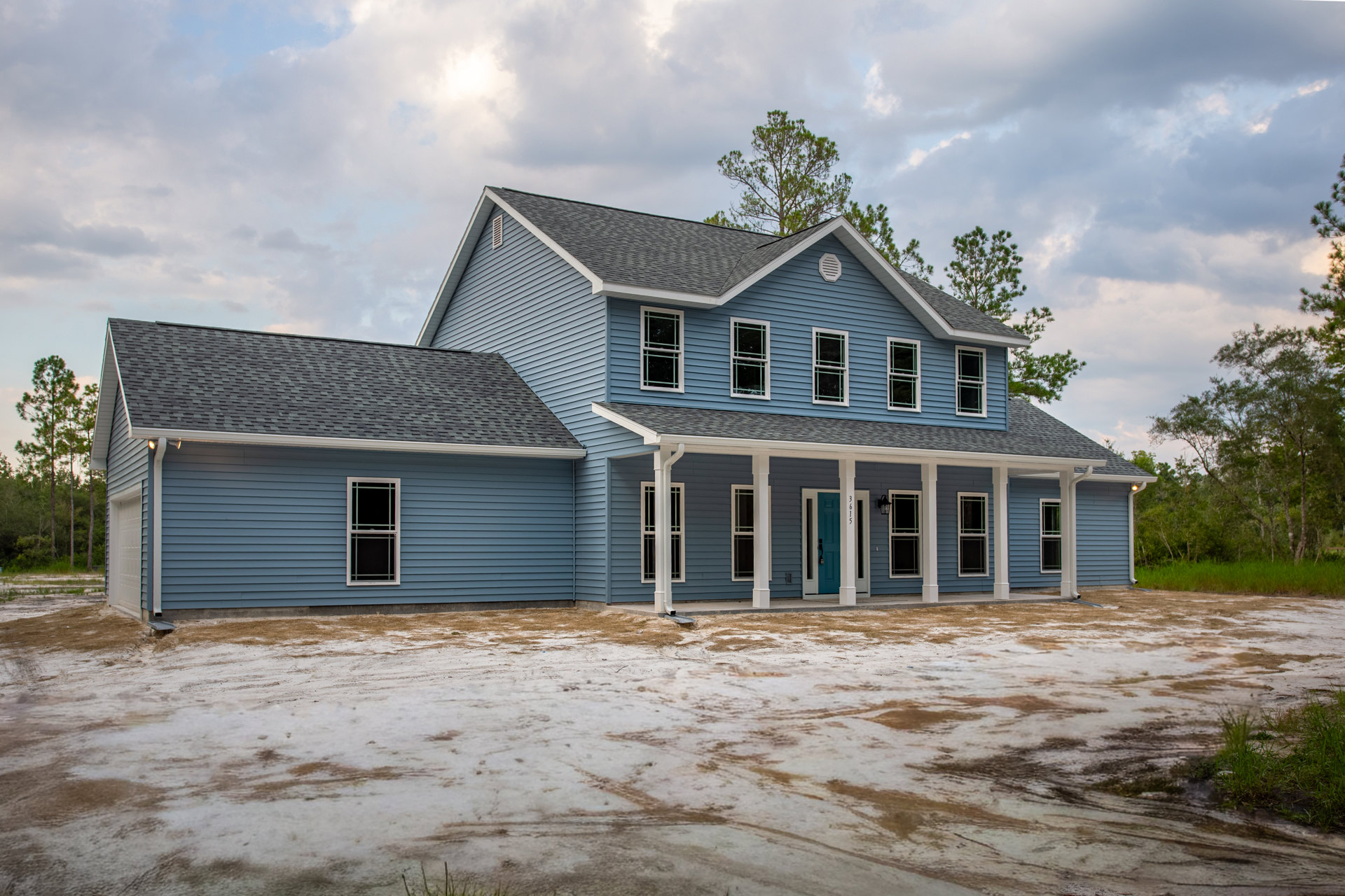Blue house with white pillars, large covered porch, multiple windows with varied frames, dirt yard in foreground, cloudy sky overhead.