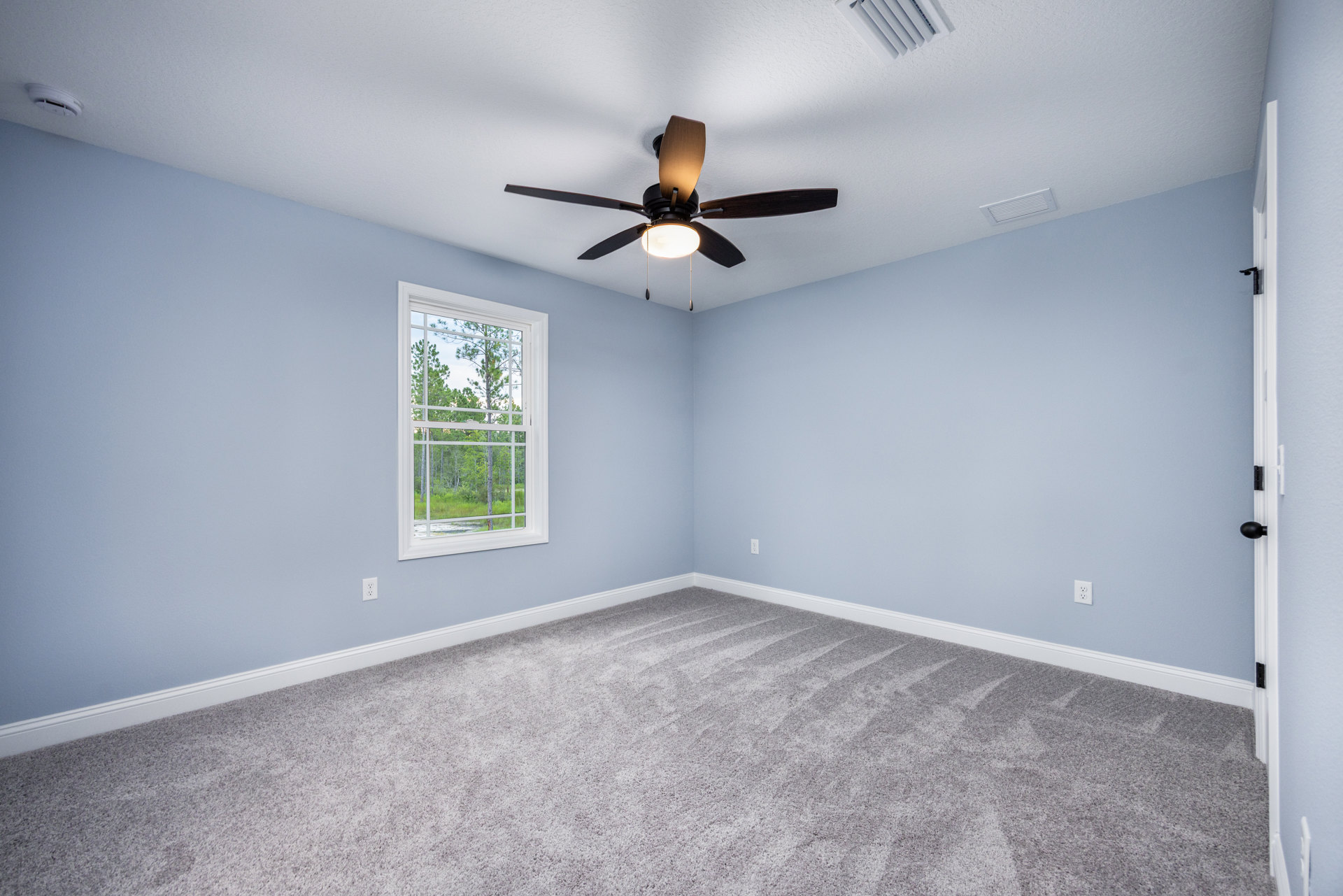 Neutral-toned carpeted room featuring a ceiling fan with light, single window overlooking green trees, white plaster walls, and visible ceiling vent.