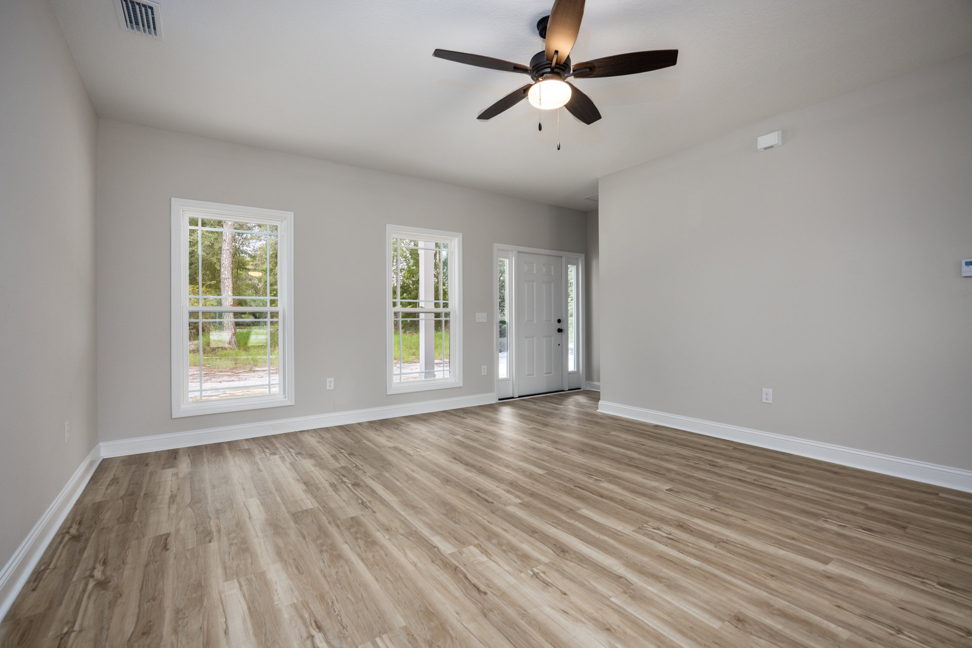 Ceiling fan with light fixture, wood flooring, white walls, white door with black knobs, large window framed in white showing green lawn and trees outside