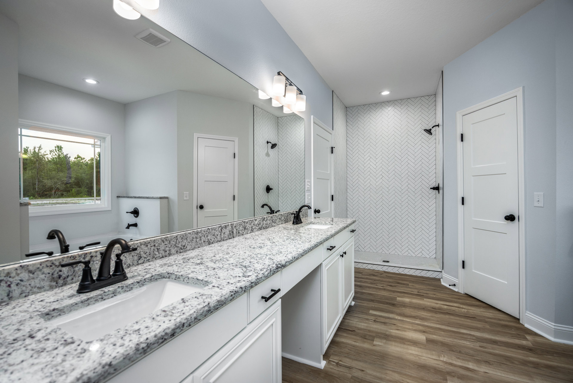Bathroom with marble countertop, black faucet, wood flooring, white door with black hardware, window showing trees outside