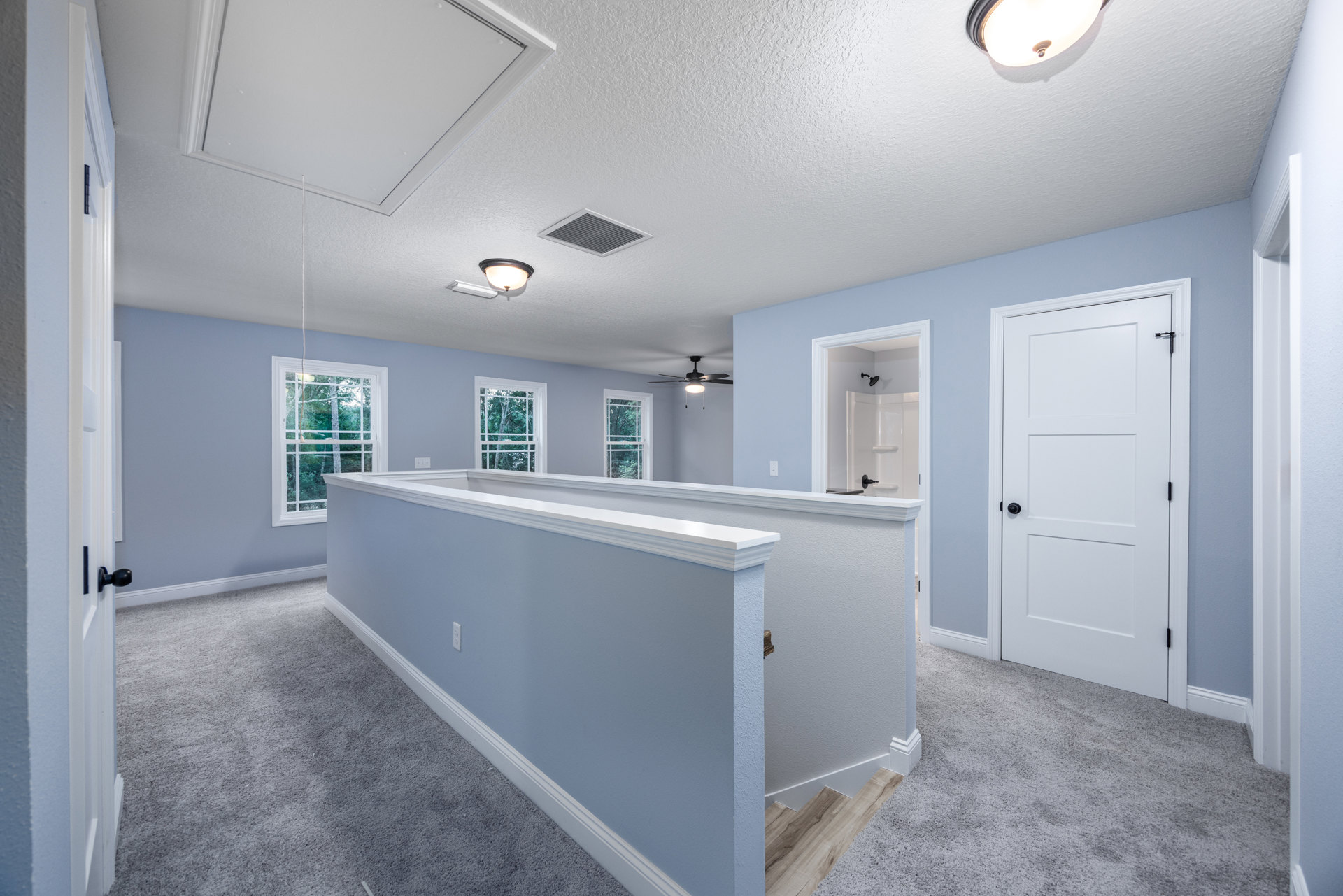 Open room with white walls, wood staircase with black railing, white door with black knob, large windows showing trees outside, tile flooring, recessed ceiling lights