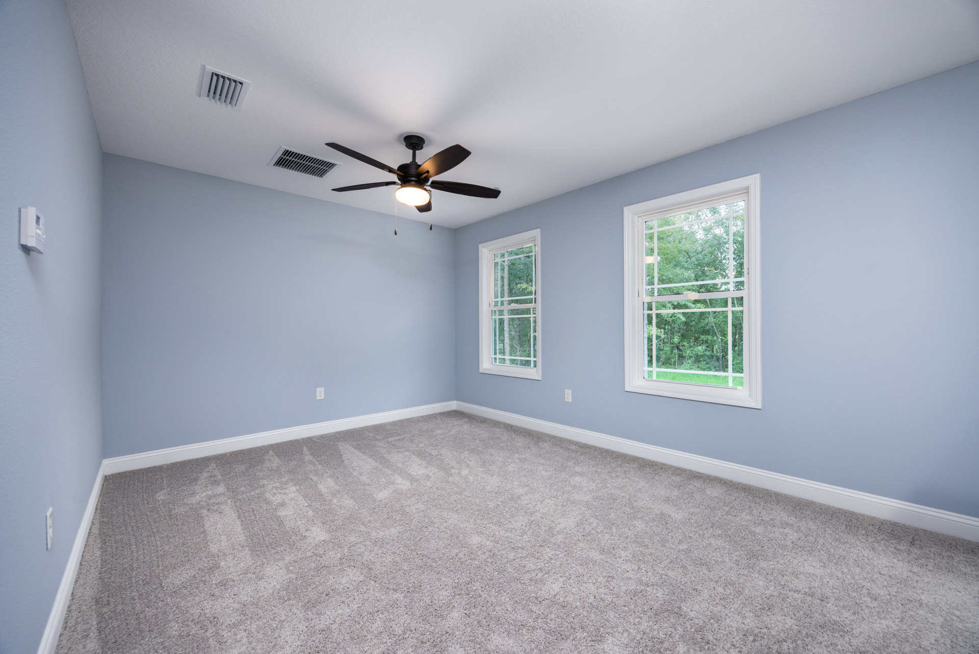 Carpeted room with blue accent wall, white trim, ceiling fan with light, two windows showing trees outside, ceiling vent