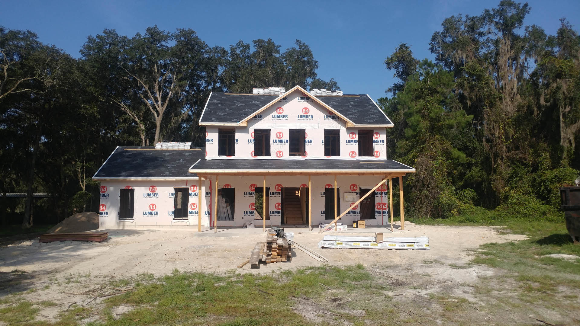 Framed house under construction with exposed wood beams, pile of lumber on dirt, trees in background, unfinished porch, person ascending temporary staircase, door displaying red