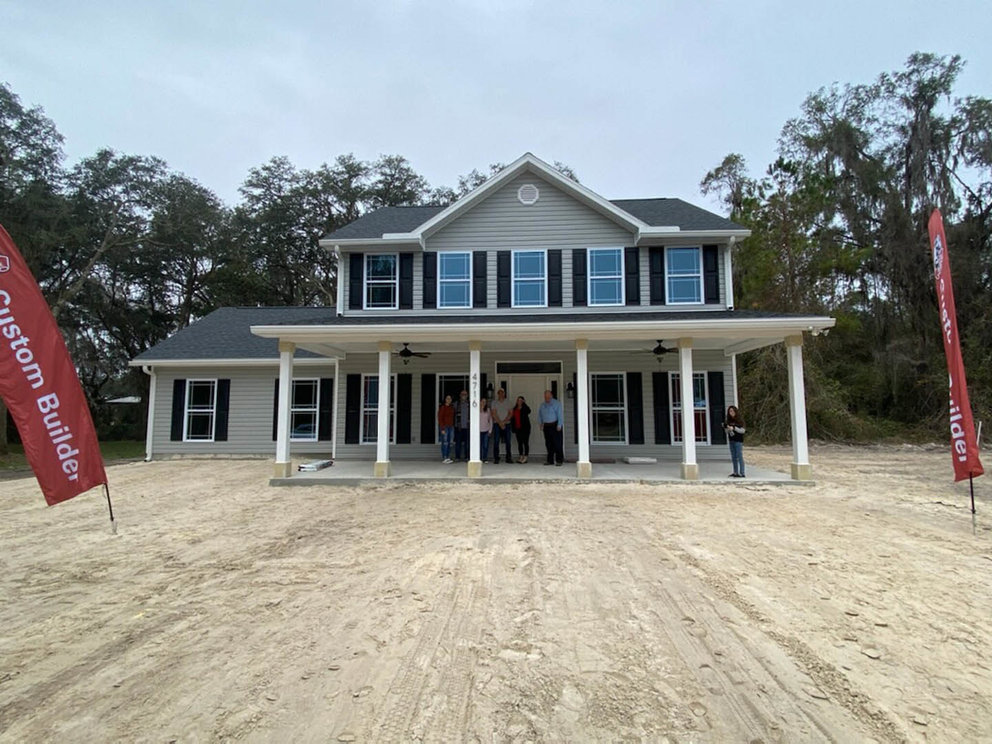 Group of people standing on dirt in front of a modern home with white framed windows, white door, and red flag featuring a white design; porch and exterior surrounded by trees