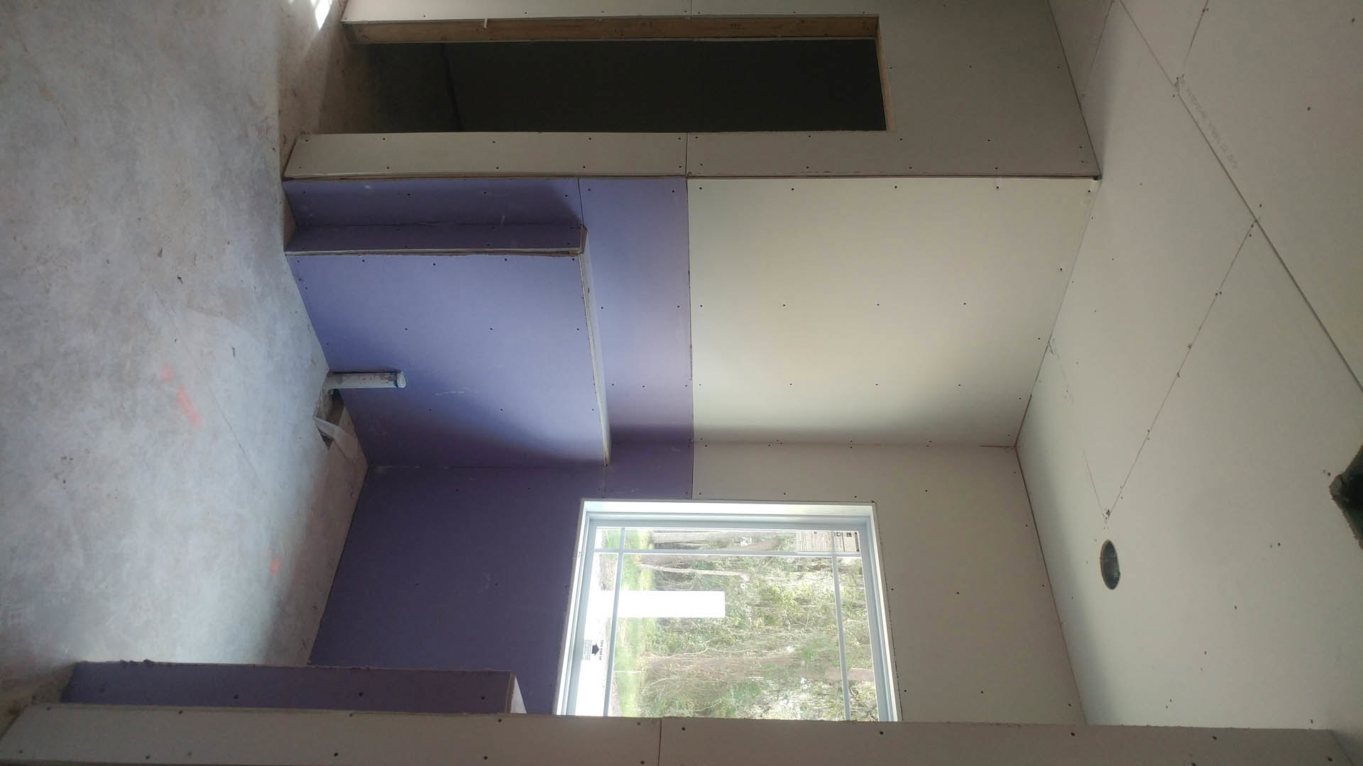 White plaster walls, window with central white rectangular object, wooden-framed blackboard, door, white floor with red mark, daylight filtering into room.