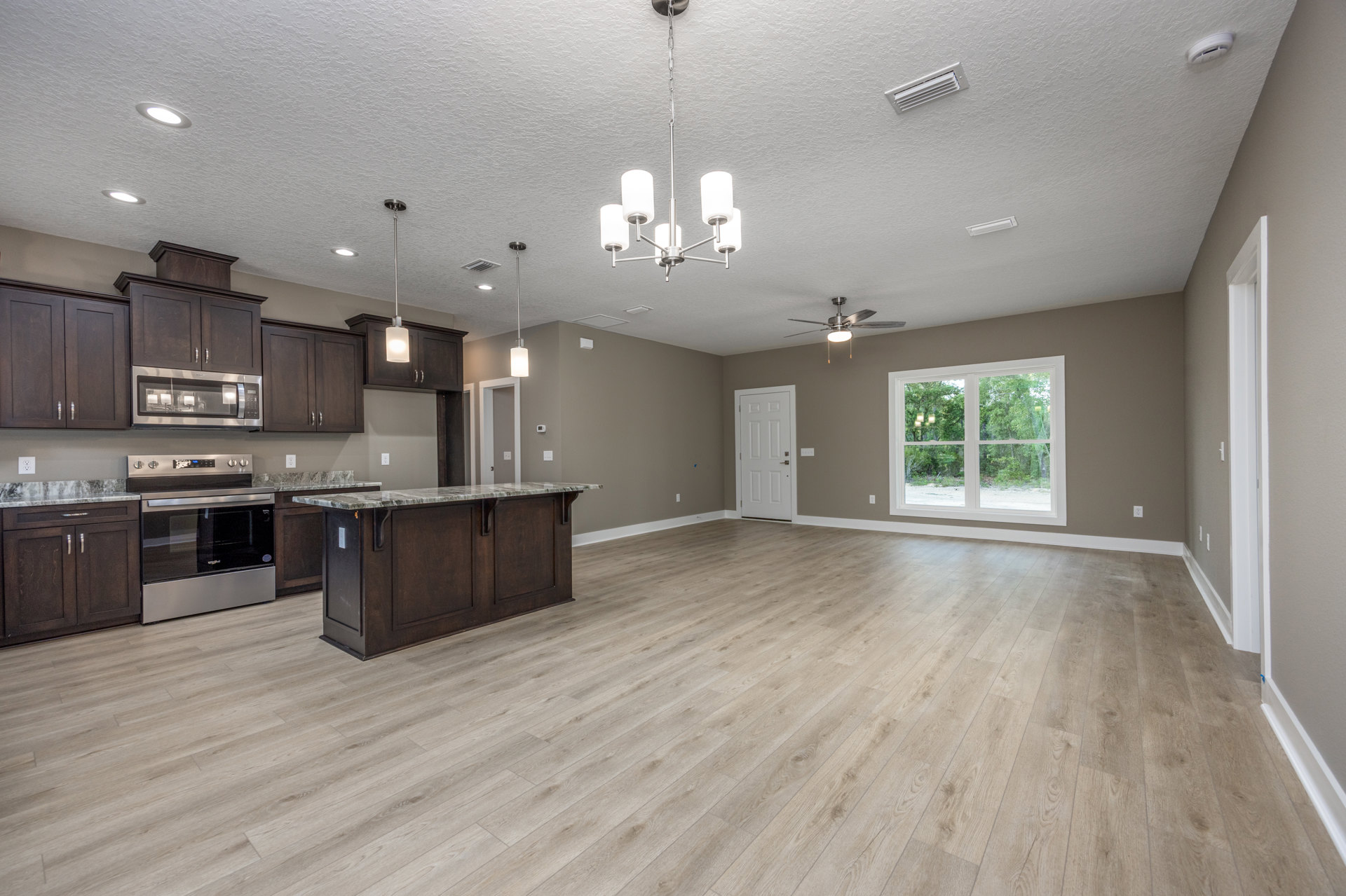 Spacious open-concept kitchen with central island, hardwood flooring, stainless steel stove with glass door, white cabinetry, large window overlooking trees, white door with silver