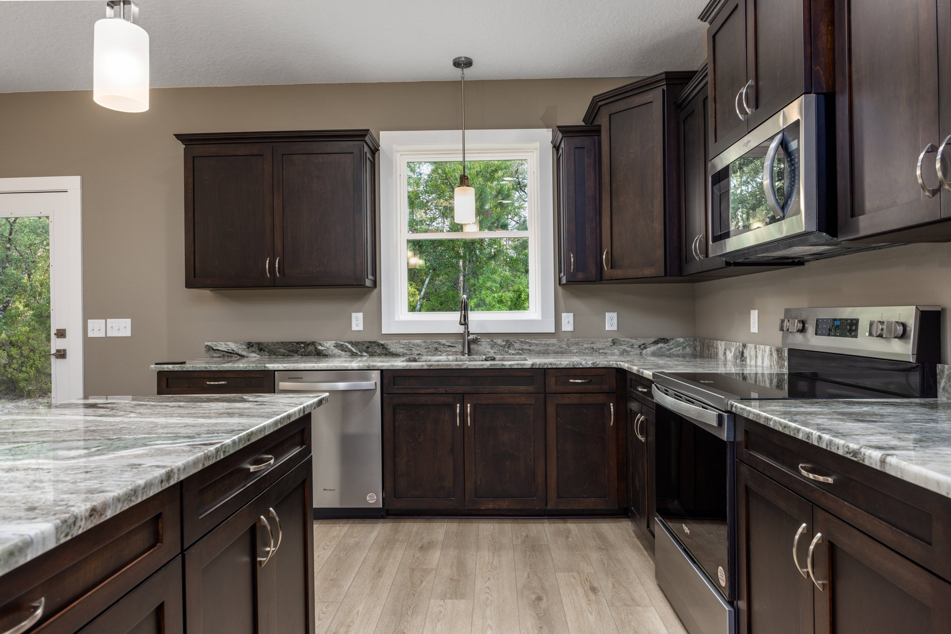 Kitchen with dark wood cabinets, silver handles, granite countertops, stainless steel microwave, white pendant light, and window providing natural light