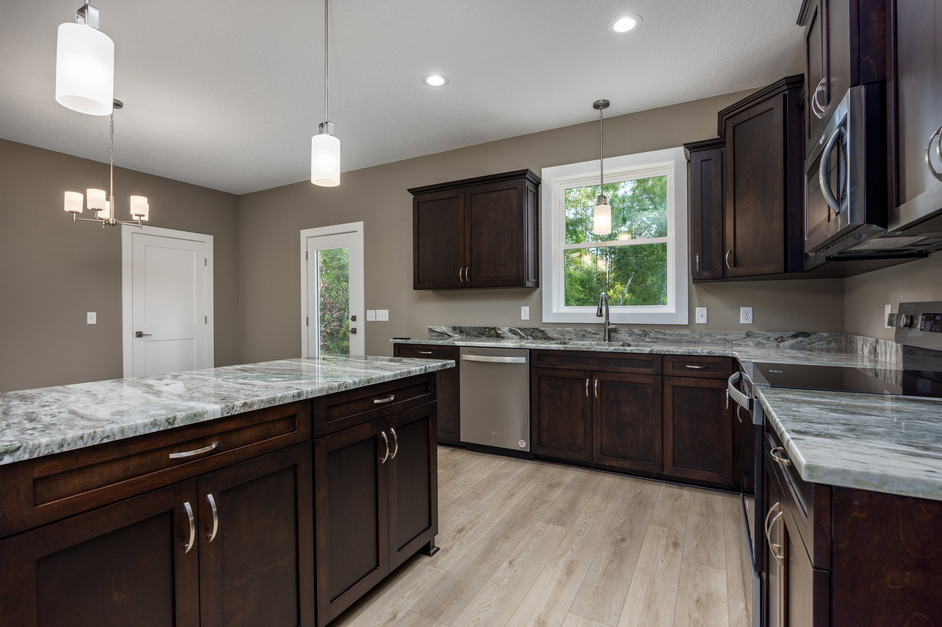 Kitchen with dark wood cabinets, marble countertops, stainless steel sink beneath a window showing trees, white door with silver handle, ceiling light fixture, and white appliance
