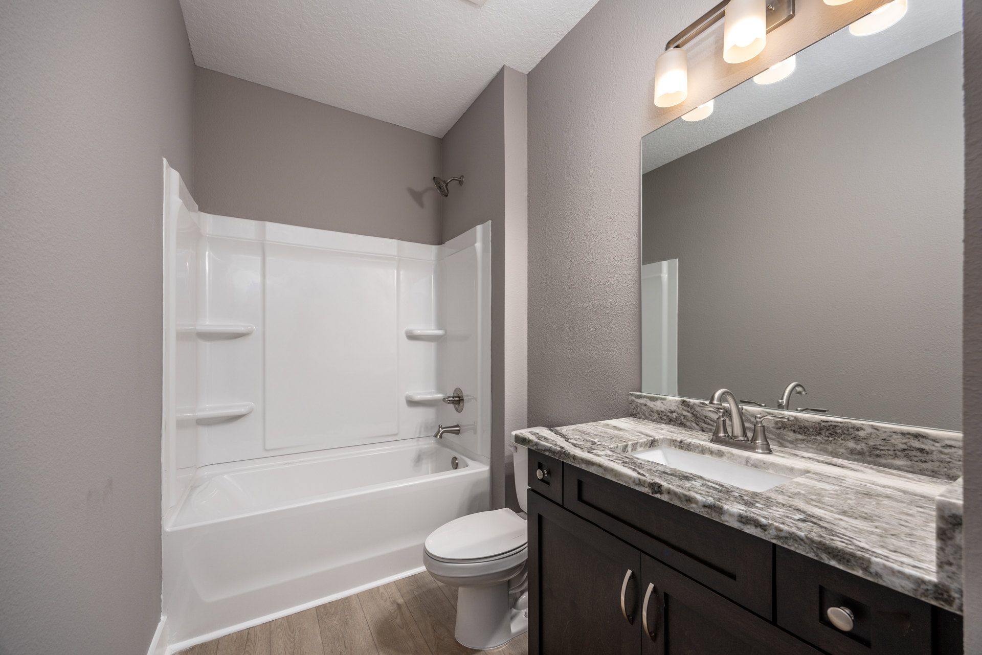 White bathroom with tile floor, white toilet, rectangular sink set in a wood cabinet with metal handles, chrome faucet, and partial view of a white bathtub.
