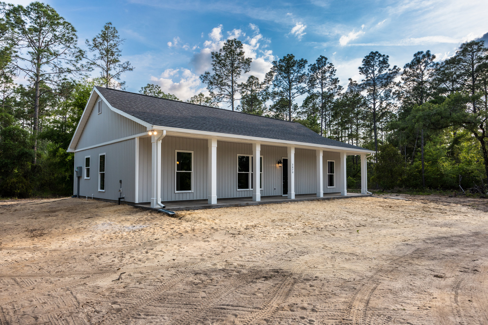 White house with white pillars, porch, and multiple windows, dirt area with tire tracks in foreground, trees and cloudy sky in background