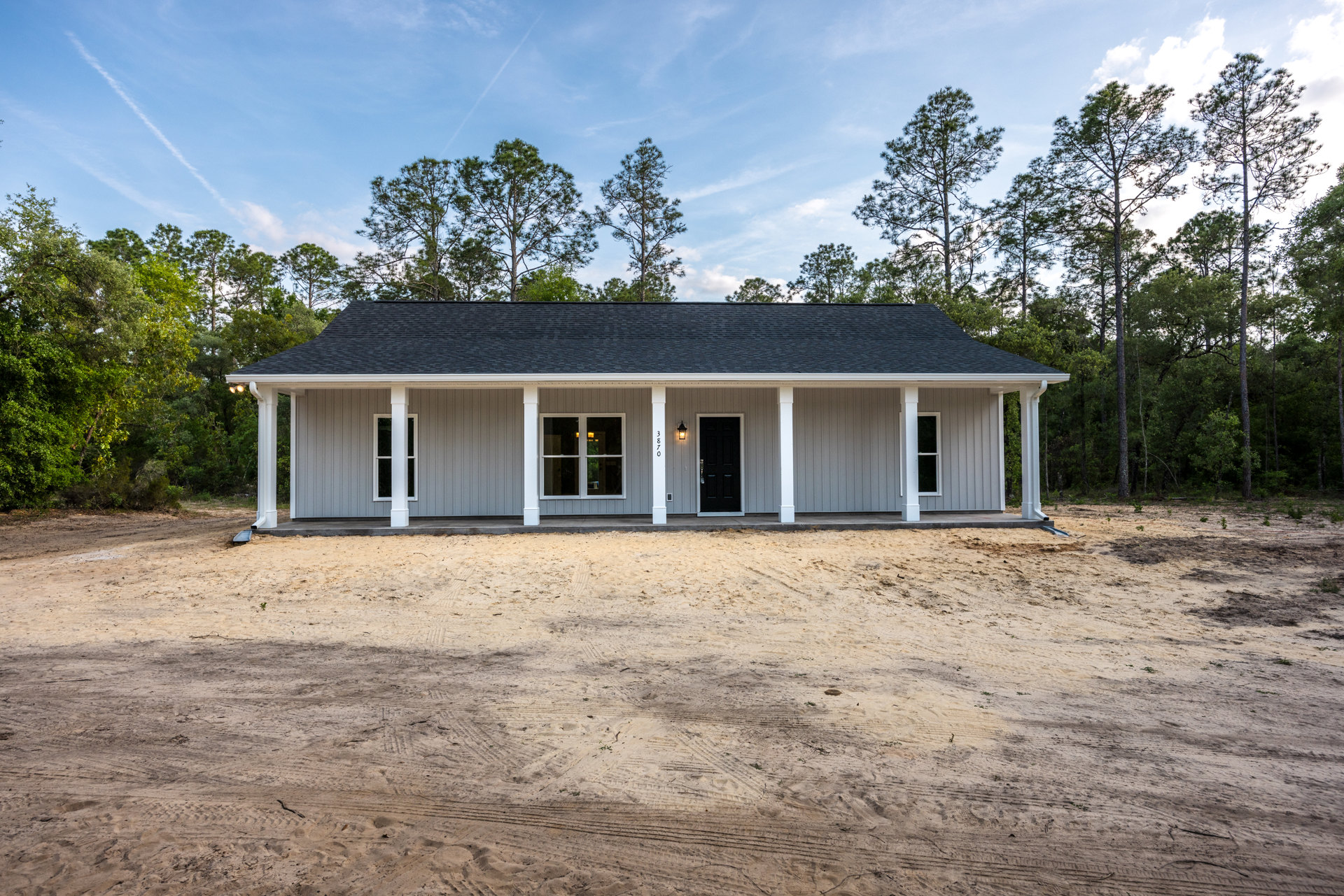 Modern home exterior with white siding, black front door, white-framed windows, dark shingle roof, surrounded by dirt lot with tire tracks and mature trees under cloudy sky