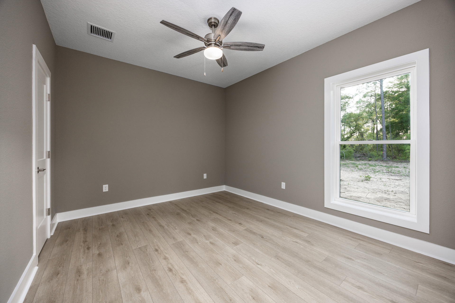 Ceiling fan with light fixture above wood flooring, window overlooking trees, white plaster walls, wall vent visible
