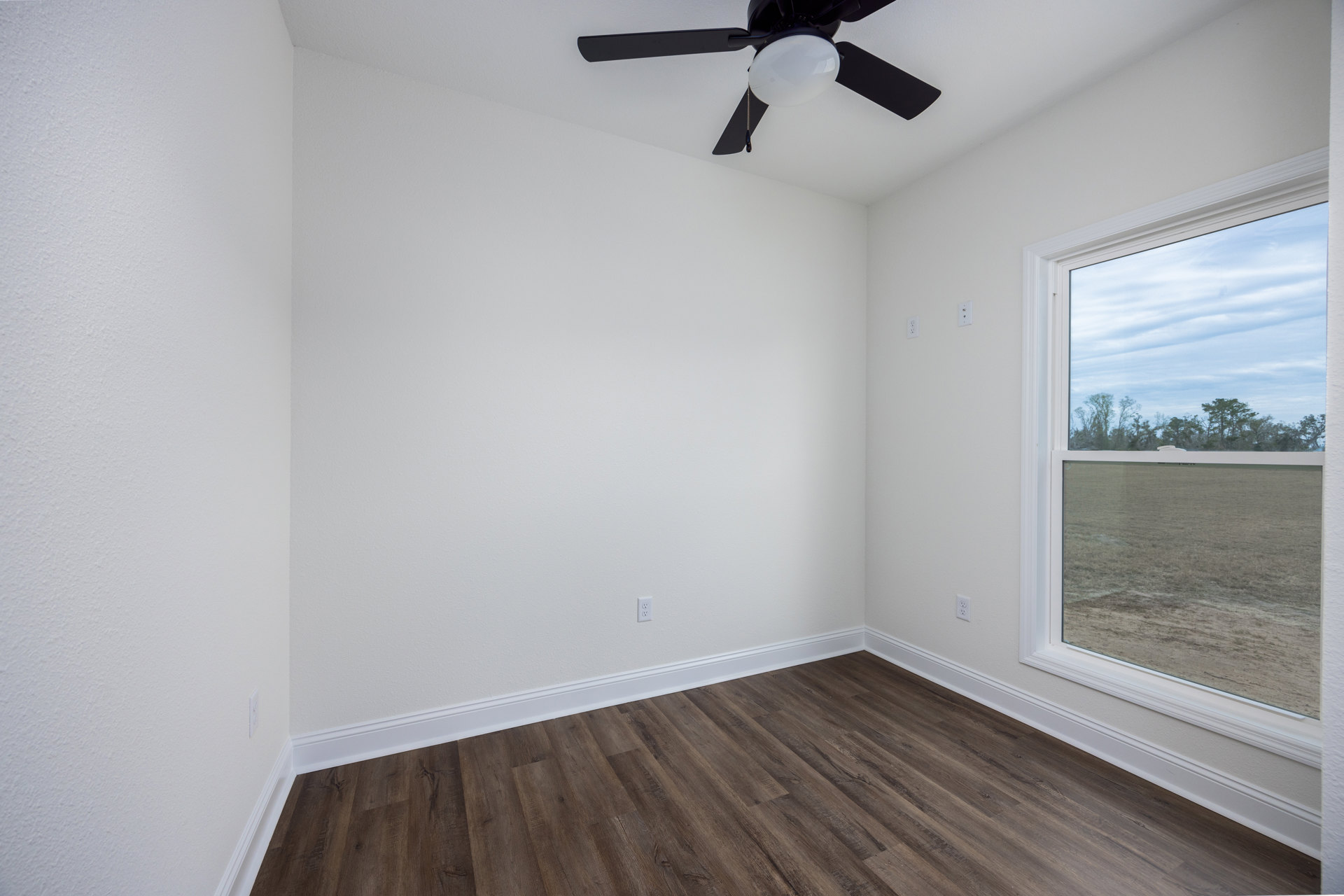 Wood floor with white baseboards, ceiling fan with light fixture, large window overlooking field and trees, smooth plaster walls