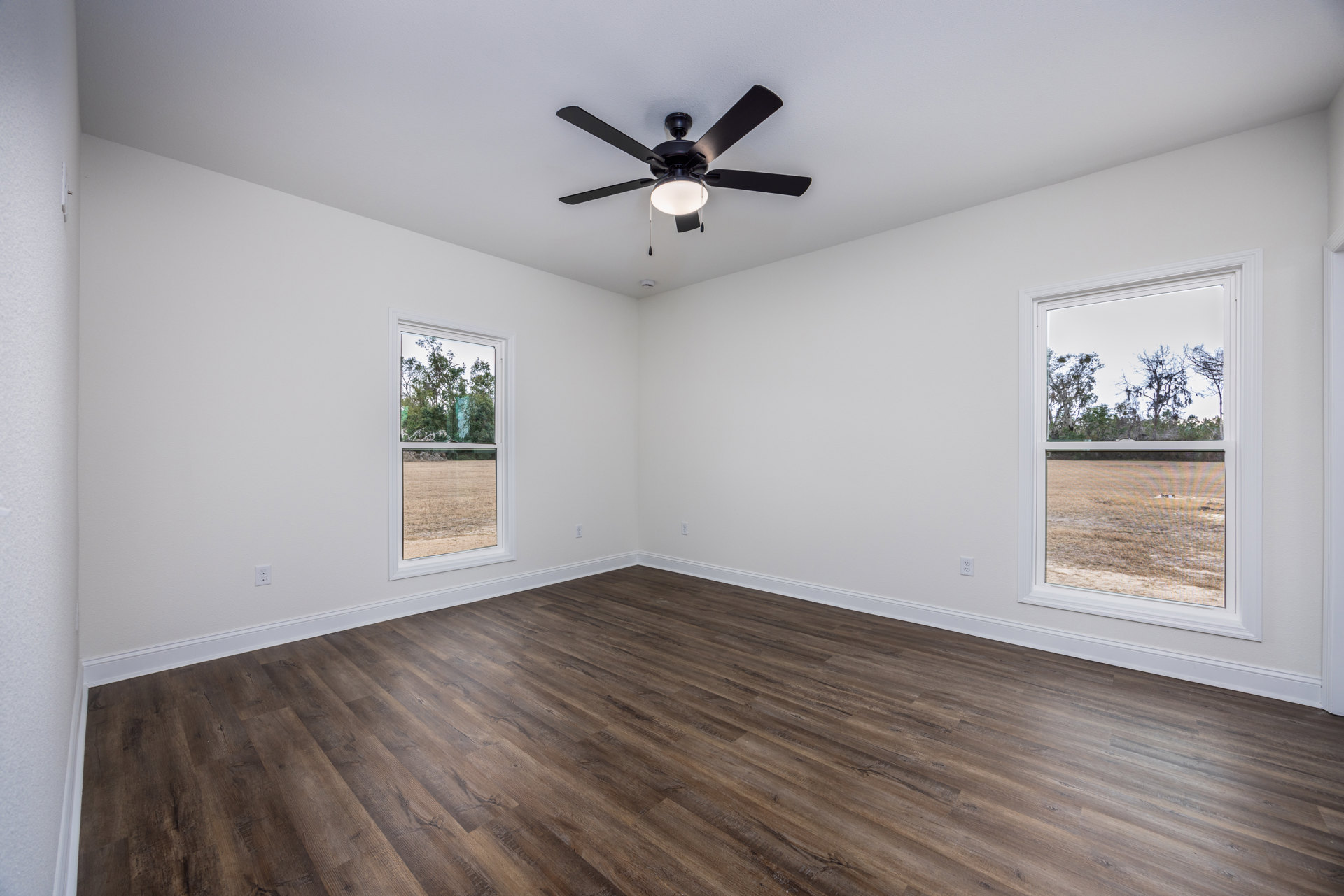 Ceiling fan with light fixture, wood flooring, white walls, multiple windows showing views of trees and open field