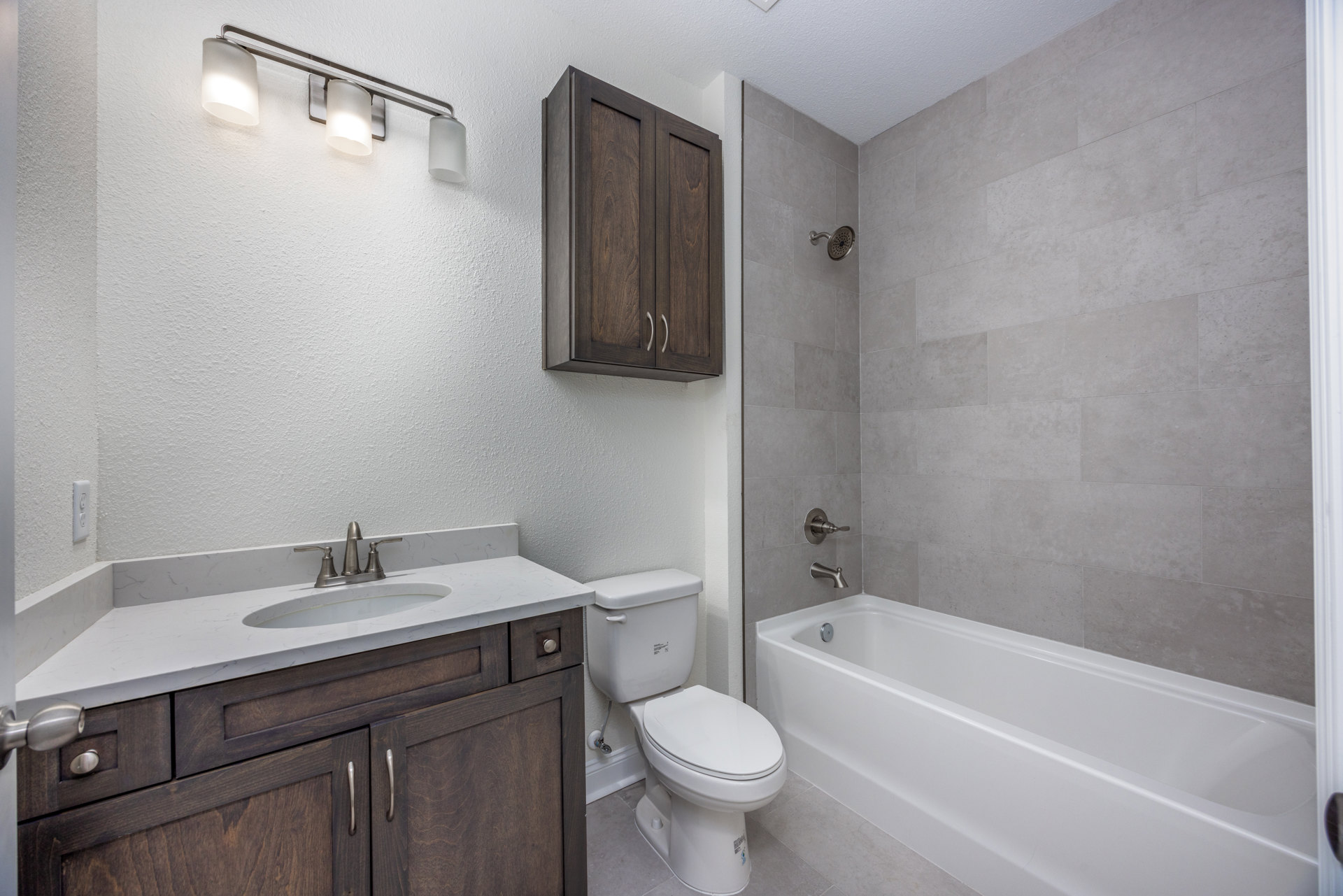 Bathroom with white tile walls, freestanding white bathtub, white toilet, wooden vanity cabinet, modern sink, chrome faucet, and overhead light fixture.