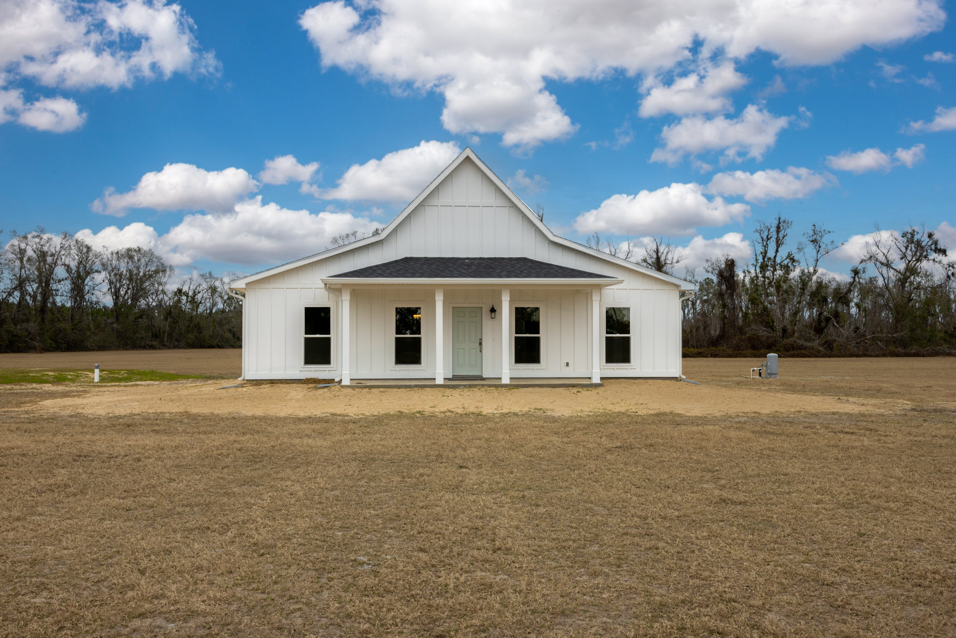 White farmhouse with triangular roof, black screened window, and spacious grassy yard surrounded by trees under a cloudy sky