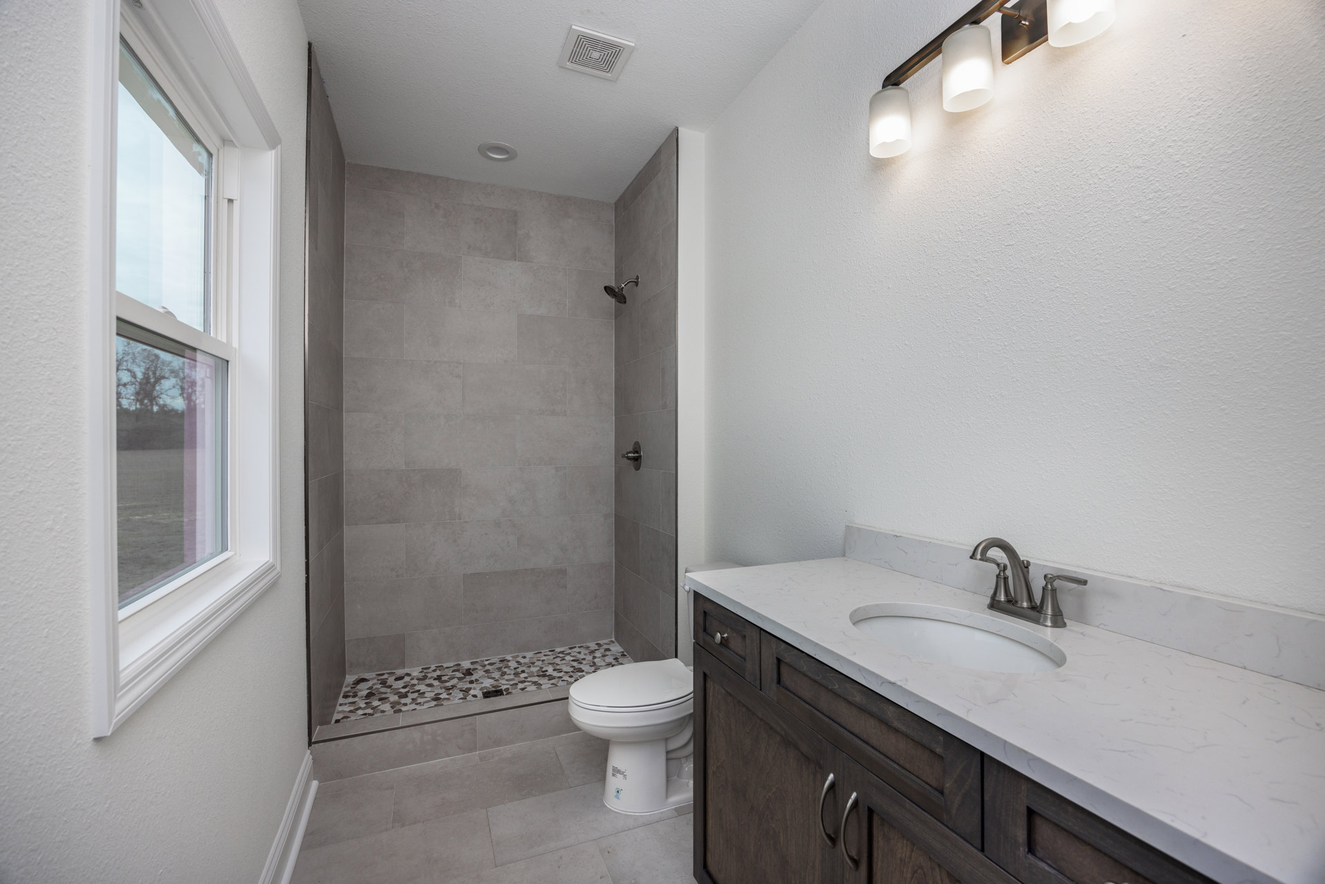 Modern bathroom with white tile walls, glass shower enclosure, white toilet with lid up, rectangular sink with chrome faucet, ceiling vent, window showing tree outside, and