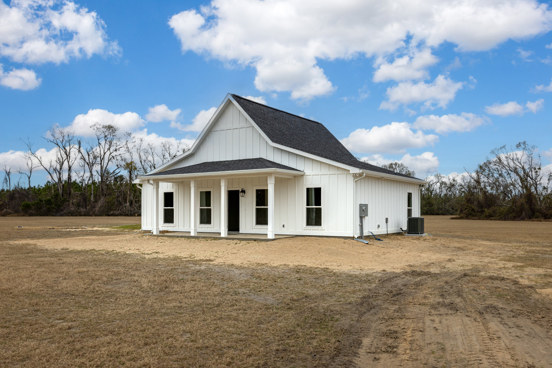 White siding house with black shingle roof, white-framed windows, dirt patch yard, black metal utility box on concrete base, surrounded by trees under blue sky
