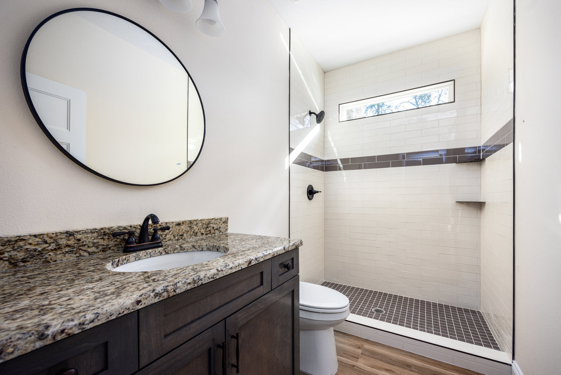 Bathroom with marble countertop vanity, undermount sink, wall-mounted mirror, glass-enclosed shower, white toilet, rectangular window showing trees, and light tile flooring.