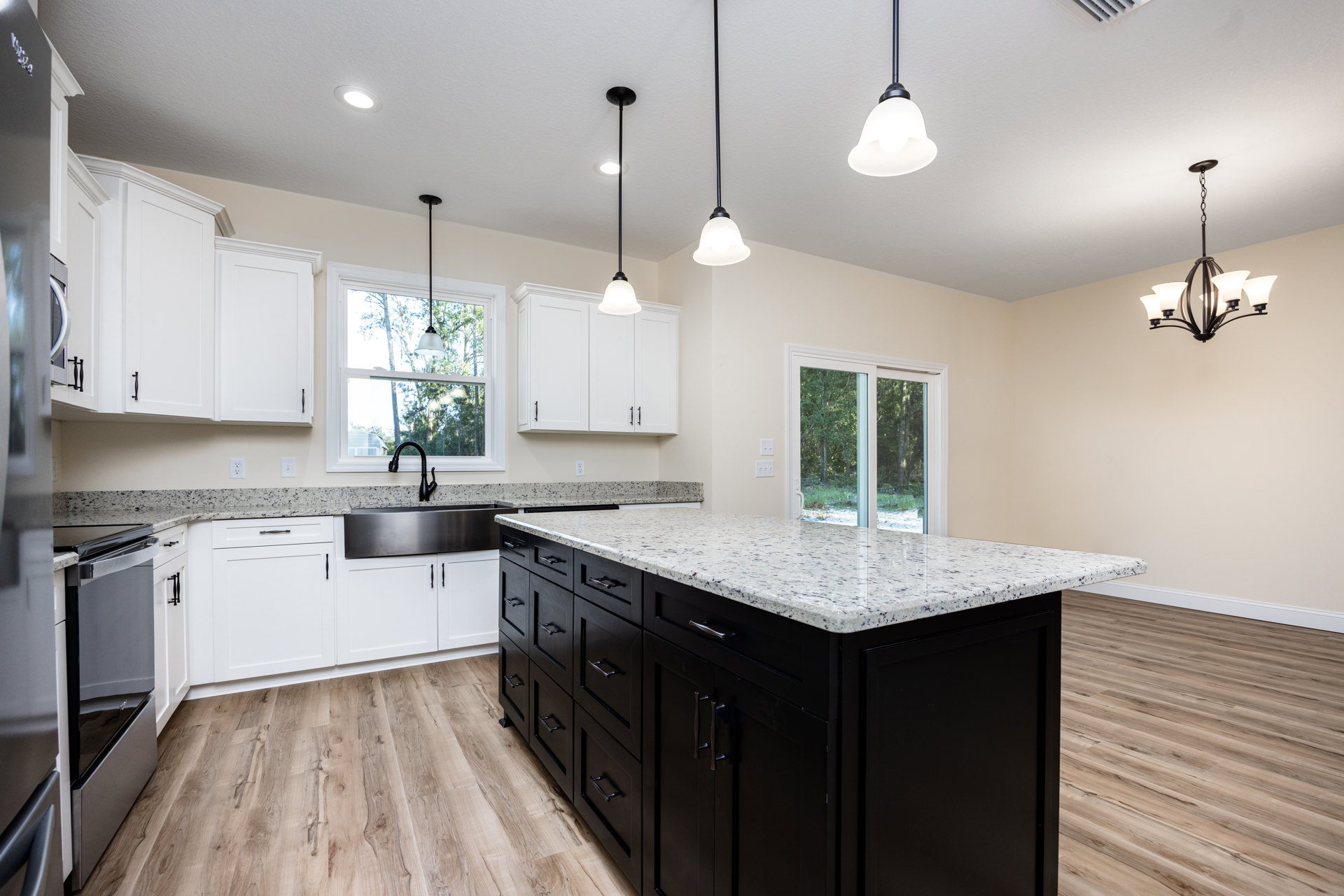 Spacious kitchen featuring a large central island with black cabinetry, white countertops, stainless steel sink, modern appliances, and windows overlooking leafy trees.