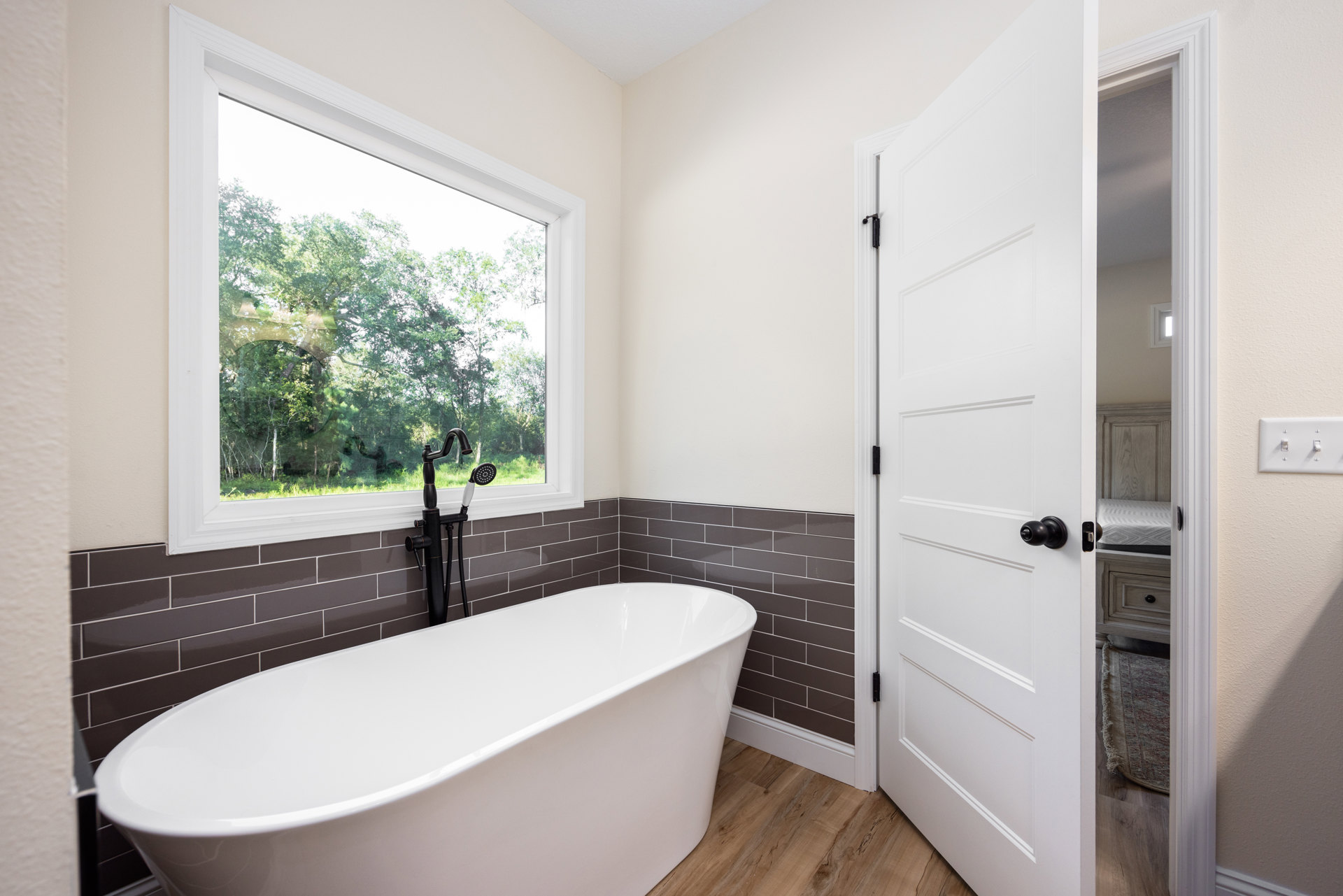 White freestanding bathtub beneath a large window, gray tile walls, chrome faucet, and wall-mounted mirror in a modern bathroom.