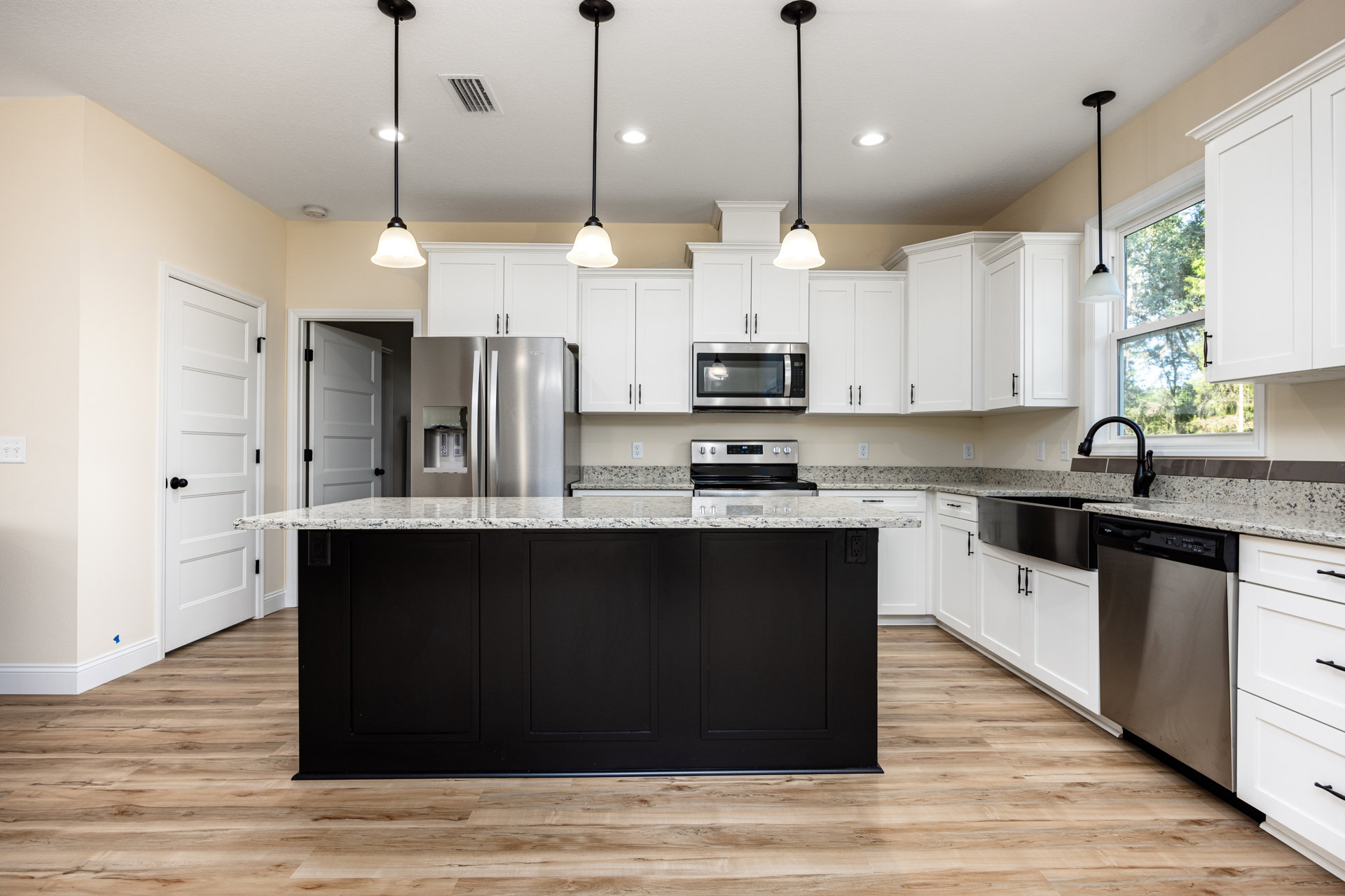 White kitchen cabinets, black marble-topped island, stainless steel refrigerator, built-in microwave, pendant light fixture, light wood flooring